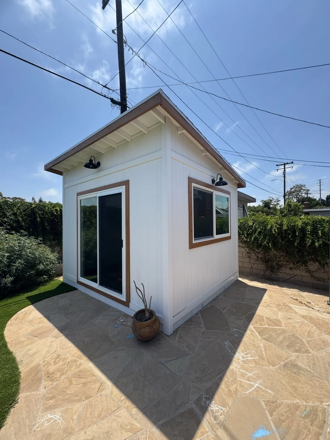 Small white house or shed with a wooden trim, large sliding glass door, and window, situated on a stone patio under clear skies."