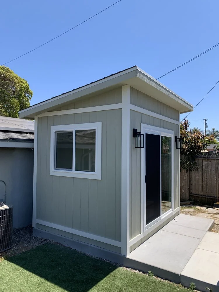 Small backyard shed with beige siding, white trim, a window, a sliding glass door, black wall-mounted light fixtures, on a concrete pad with grass and a wooden fence in the background under a clear blue sky.