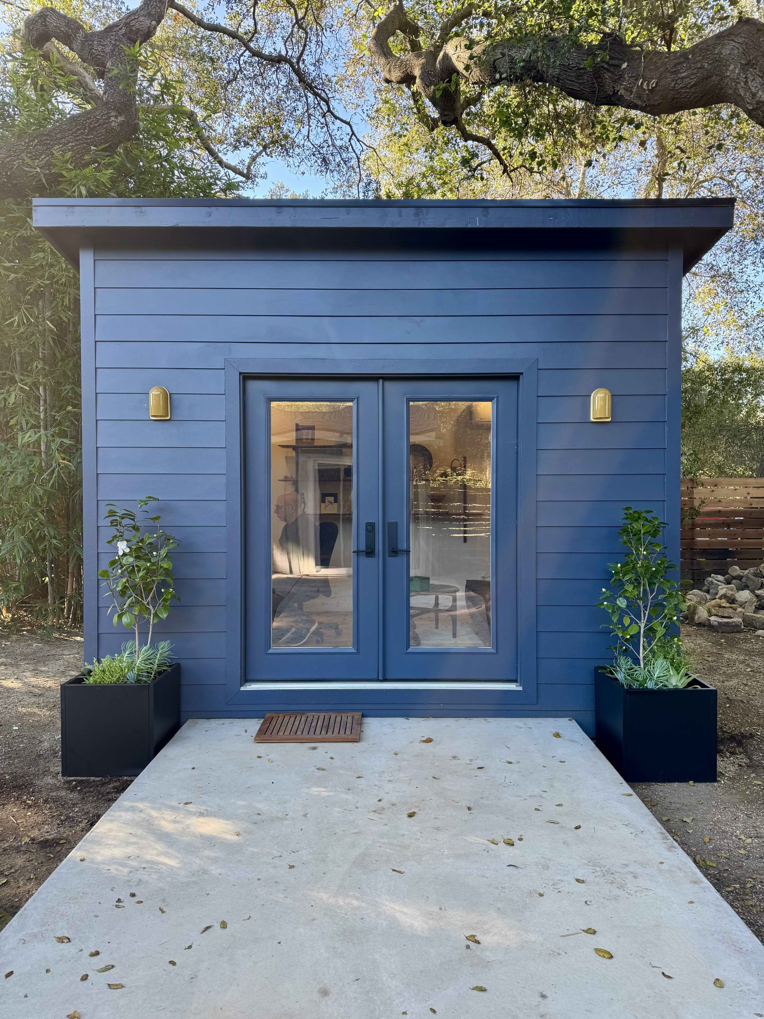 A blue studio shed with a glass double door, two yellow wall lamps on either side, and a wooden mat outside. The building is surrounded by trees and plants, with a concrete pathway leading to the entrance.