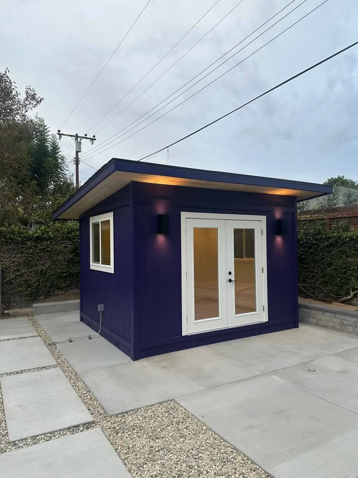 A small modern blue shed with white trim and glass doors sitting on a paved patio area, with windows and exterior lighting, surrounded by greenery and utility power lines in the background.