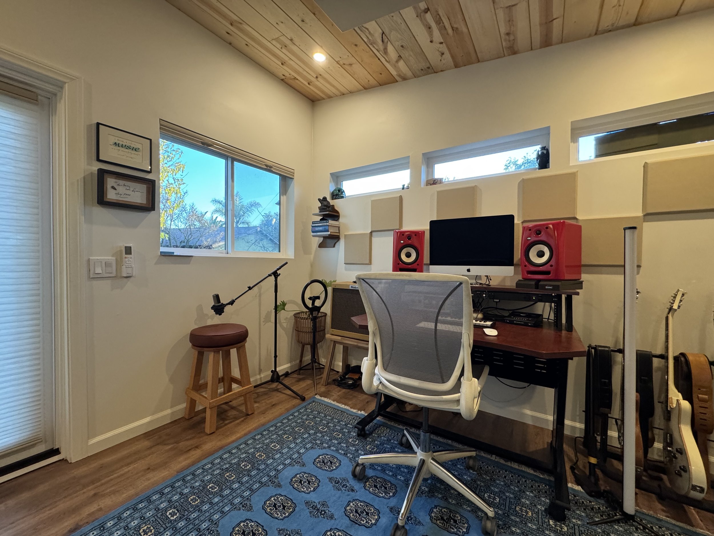 A cozy home office studio shed with a wooden ceiling, built-in shelves, multiple windows, a computer desk with an iMac, red speakers, and music equipment, a blue patterned rug, and musical instruments in a well-lit space.