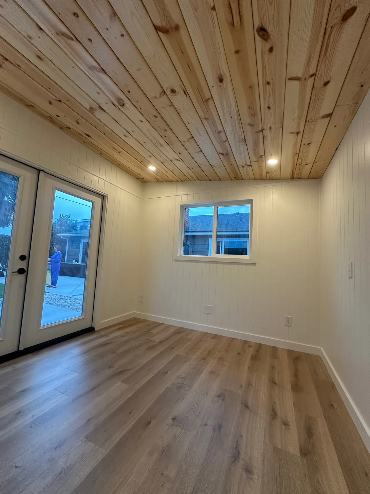 Empty room with wooden floor and ceiling, white paneled walls, a window, and a sliding glass door showing a person outside.