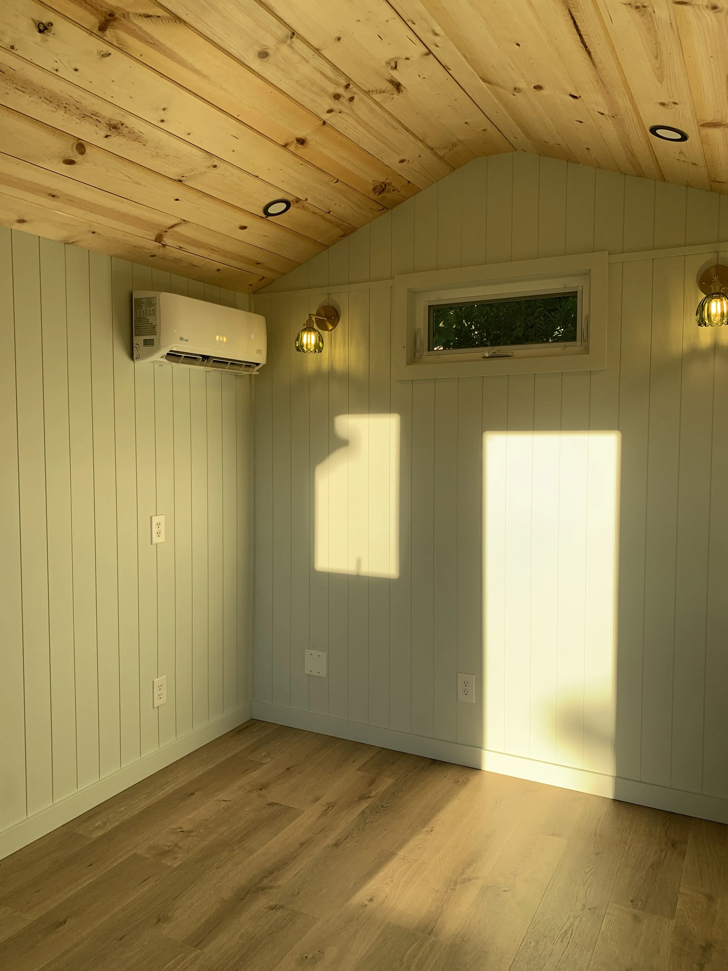 A room with wood-paneled ceiling and wall, a small window, wall-mounted lights, an air conditioner, and multiple electrical outlets.