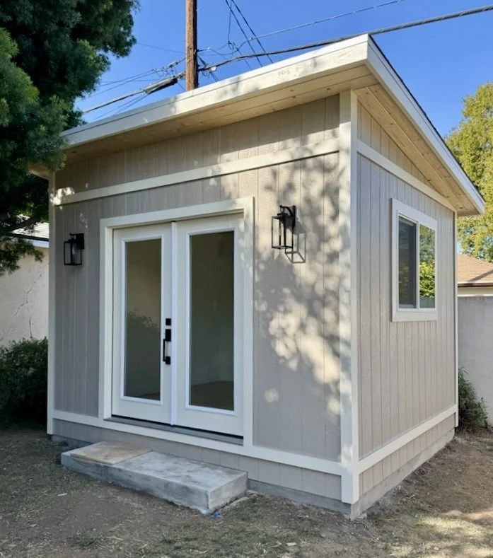 Small modern shed with sliding glass doors and exterior wall-mounted black light fixtures, surrounded by trees and a concrete step at the entrance.