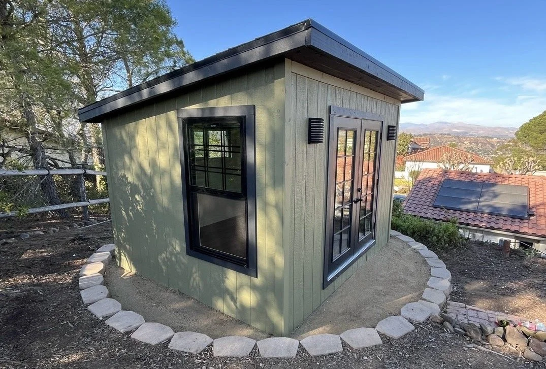 Small green outdoor shed with black-framed windows and double glass doors, surrounded by a circular border of white stones, on a hillside with oak trees and distant rooftops with solar panels.