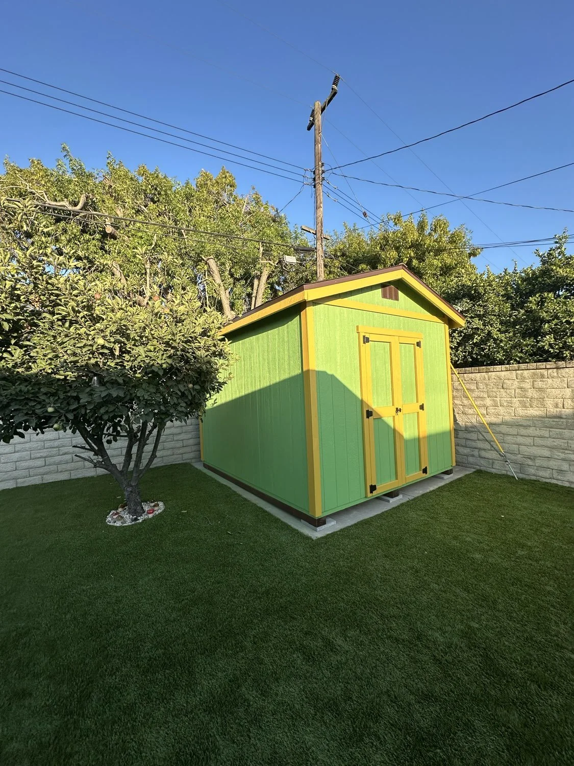 A small, colorful storage shed painted green with yellow trim located in a backyard with well-maintained grass, a tree, a brick wall, and power lines overhead.