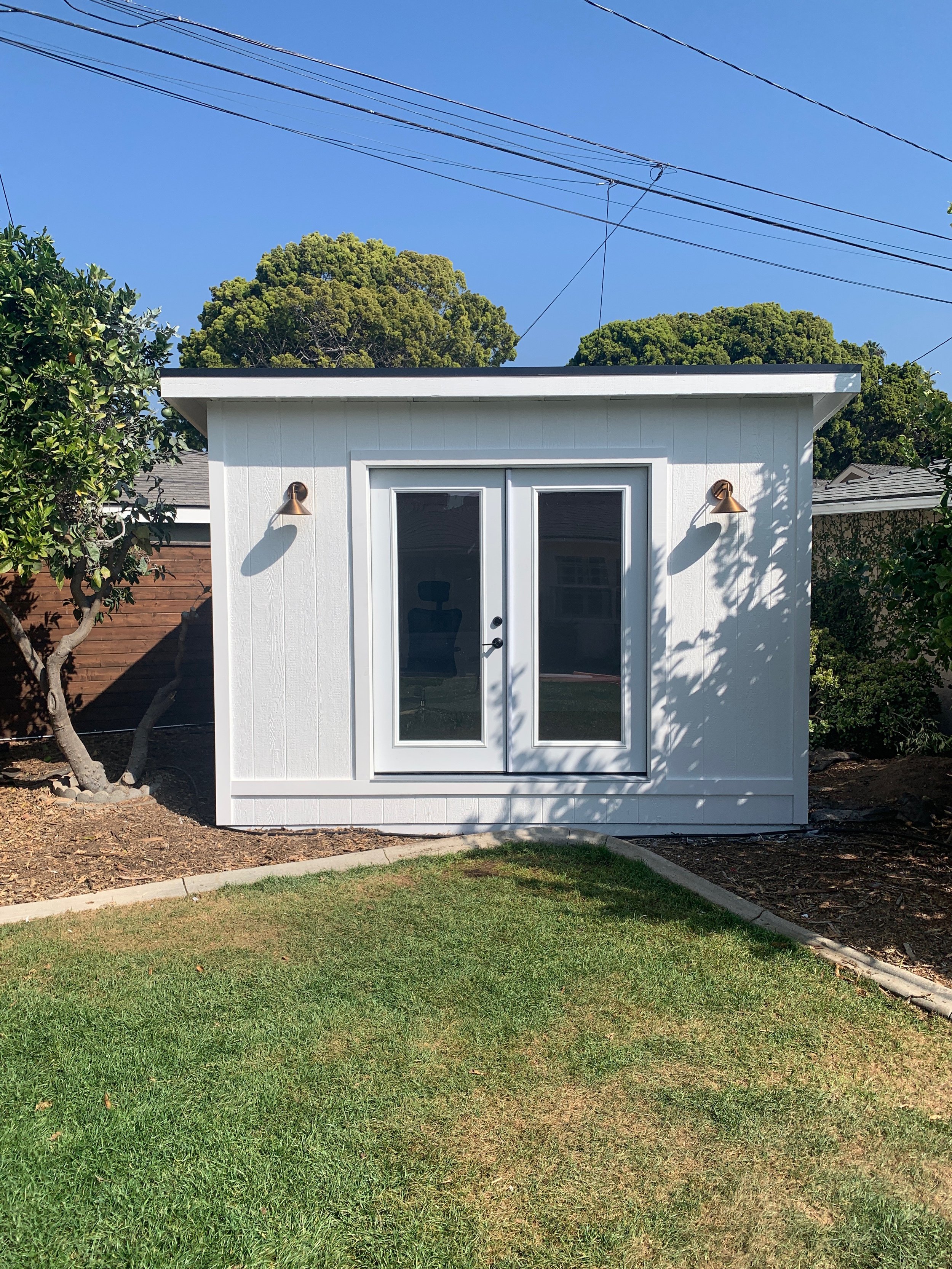 Small white shed with double glass doors, two outdoor wall-mounted lights, surrounded by greenery, with a lawn in front, under a clear blue sky, and power lines overhead.
