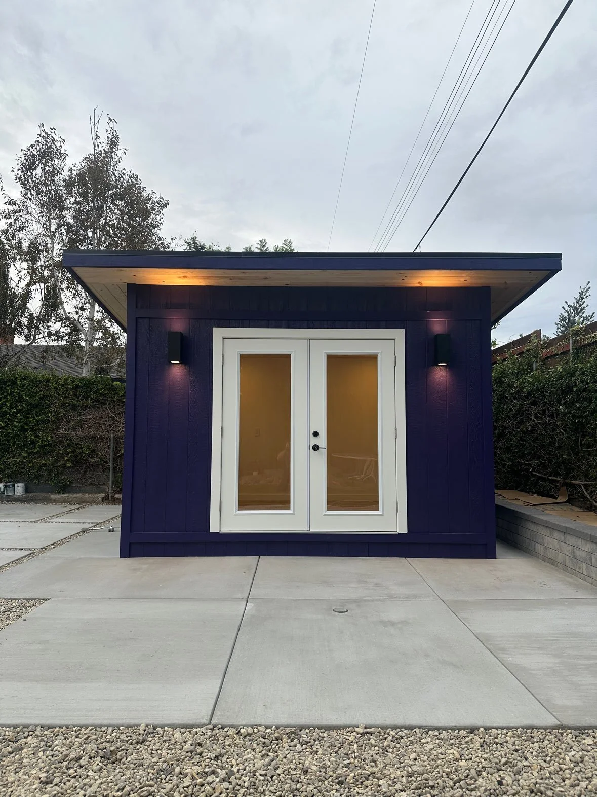 Small blue modern shed or tiny house with glass double doors, exterior wall-mounted lights, and a flat roof, situated on a concrete driveway with a gravel strip in front, surrounded by a hedge and trees.