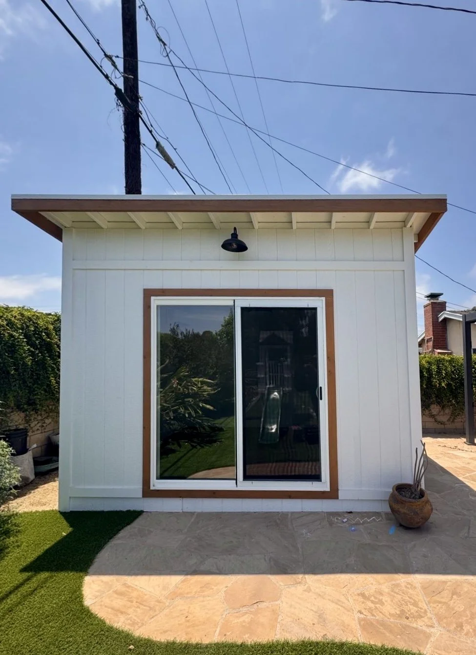 White studio shed with sliding glass door, wooden trim, black outdoor light fixture, and potted plant outside, with a backyard and utility pole with wires against a clear blue sky.
