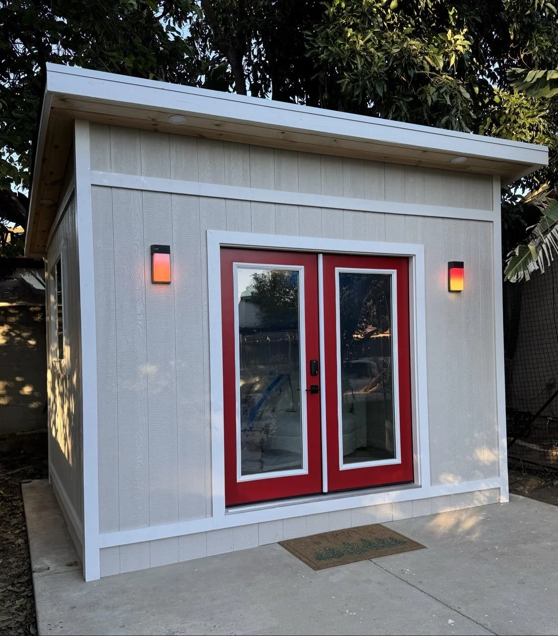 Small shed with white walls, red double doors with glass panels, and two wall-mounted lights, situated on a concrete pad with trees in the background.