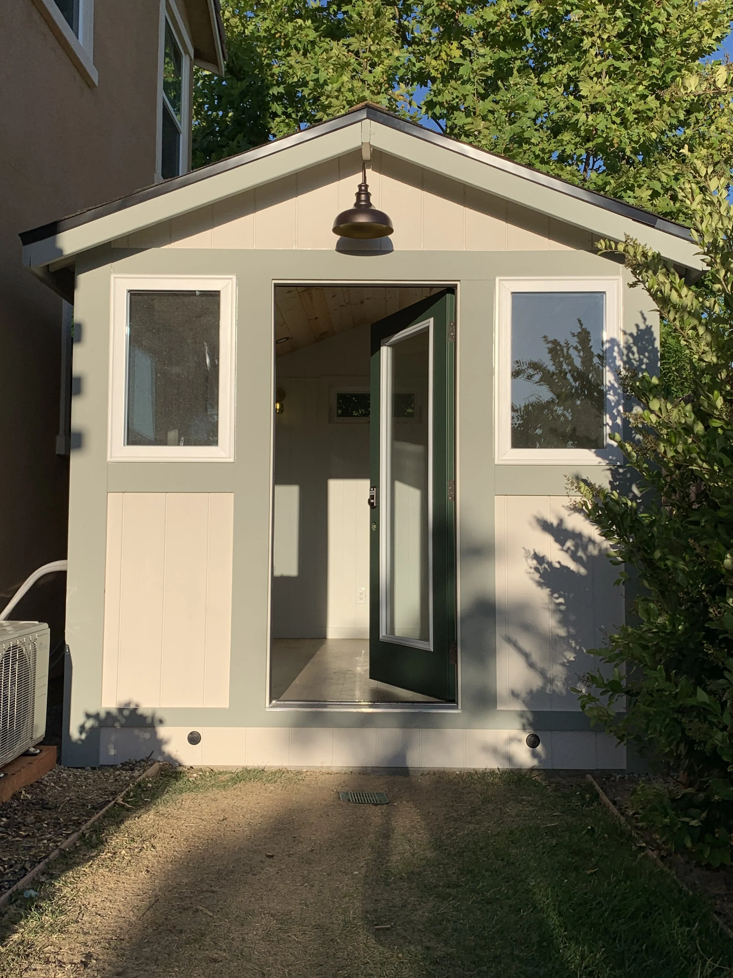 A small building or shed with a gable roof, painted in light colors, with an open door and two windows, one on each side of the door, surrounded by green trees and bushes. The shed is situated on a gravel pathway with some grass and an air conditioni