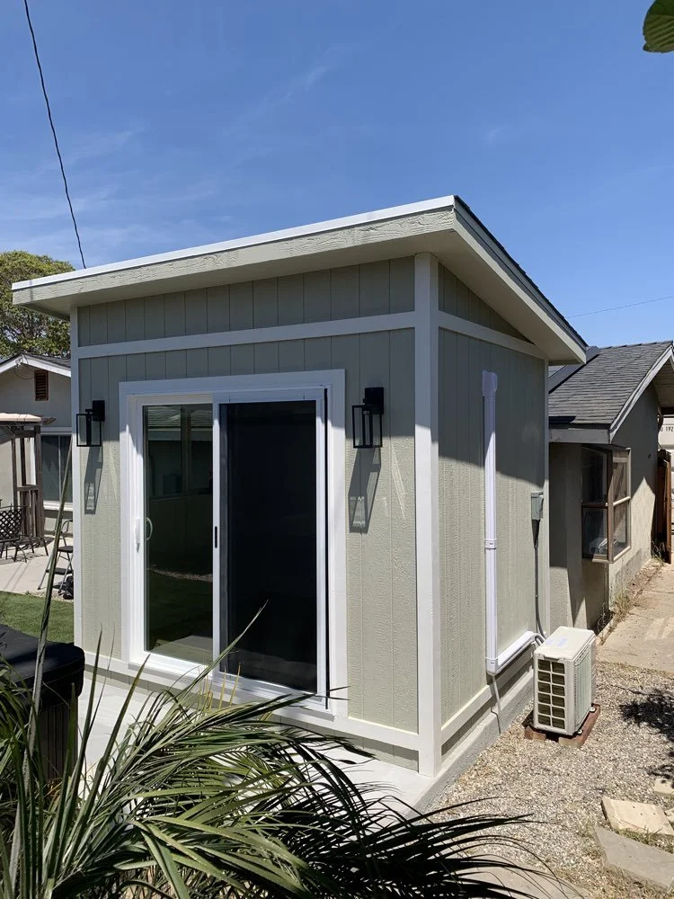 Backyard shed with gray siding, white trim, and a sliding glass door, with outdoor wall lamps, and an air conditioning unit outside.