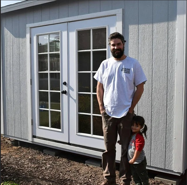 A man and a child standing in front of a gray shed with double glass doors.