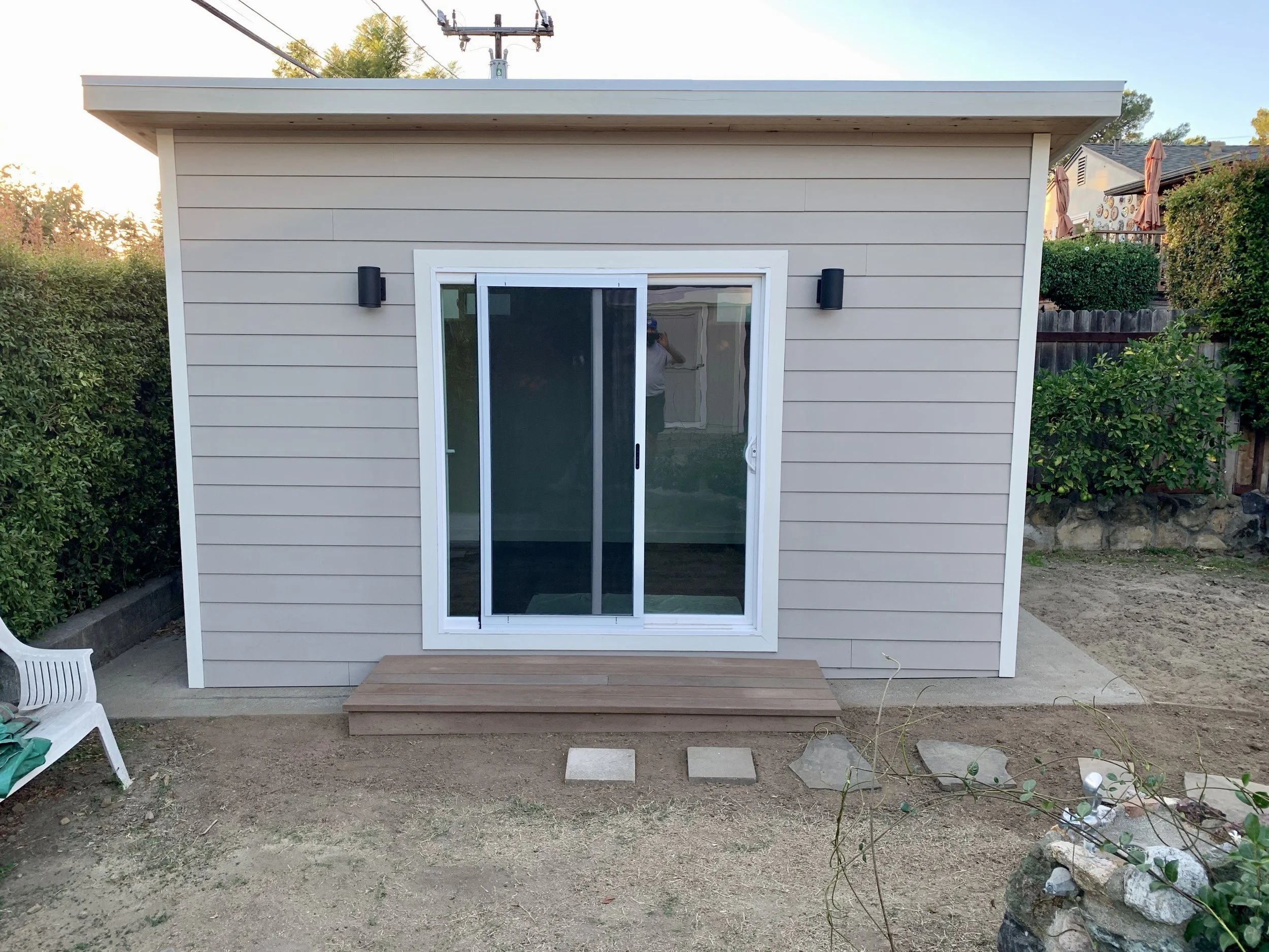 Small beige shed with sliding glass door and black wall-mounted lights, set on a concrete pad with a small wooden step in a backyard with bushes, trees, and neighboring houses.
