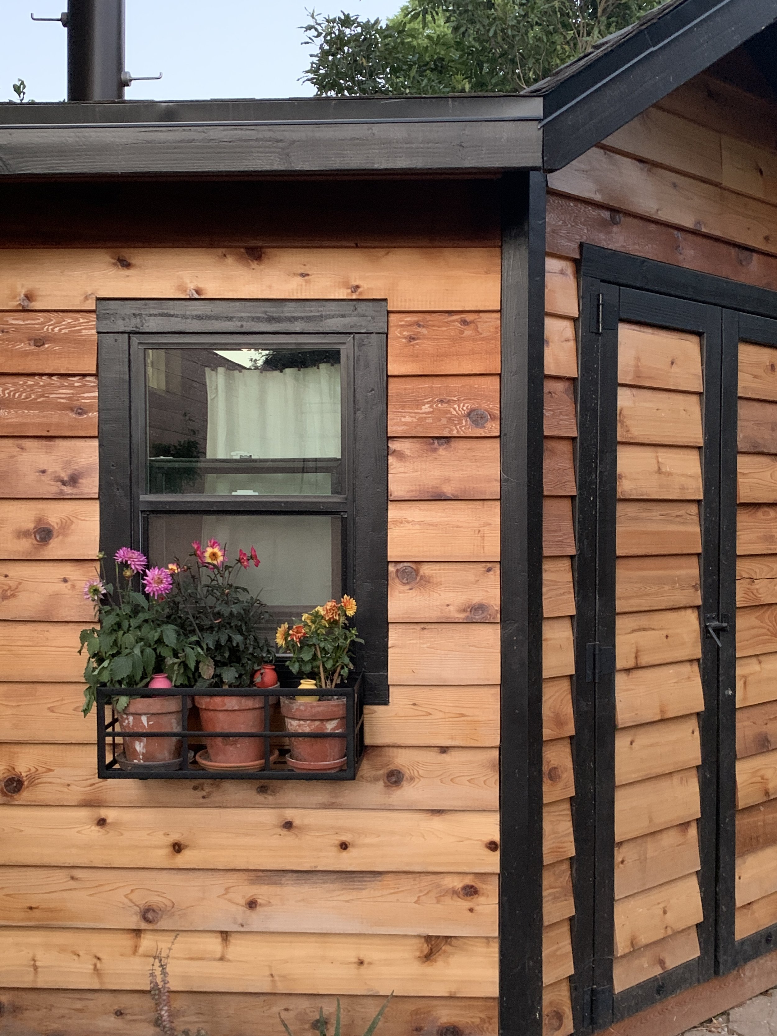 Close-up of a wooden shed exterior with a black-framed window and a small flower box with pink and yellow flowers on a black metal stand.
