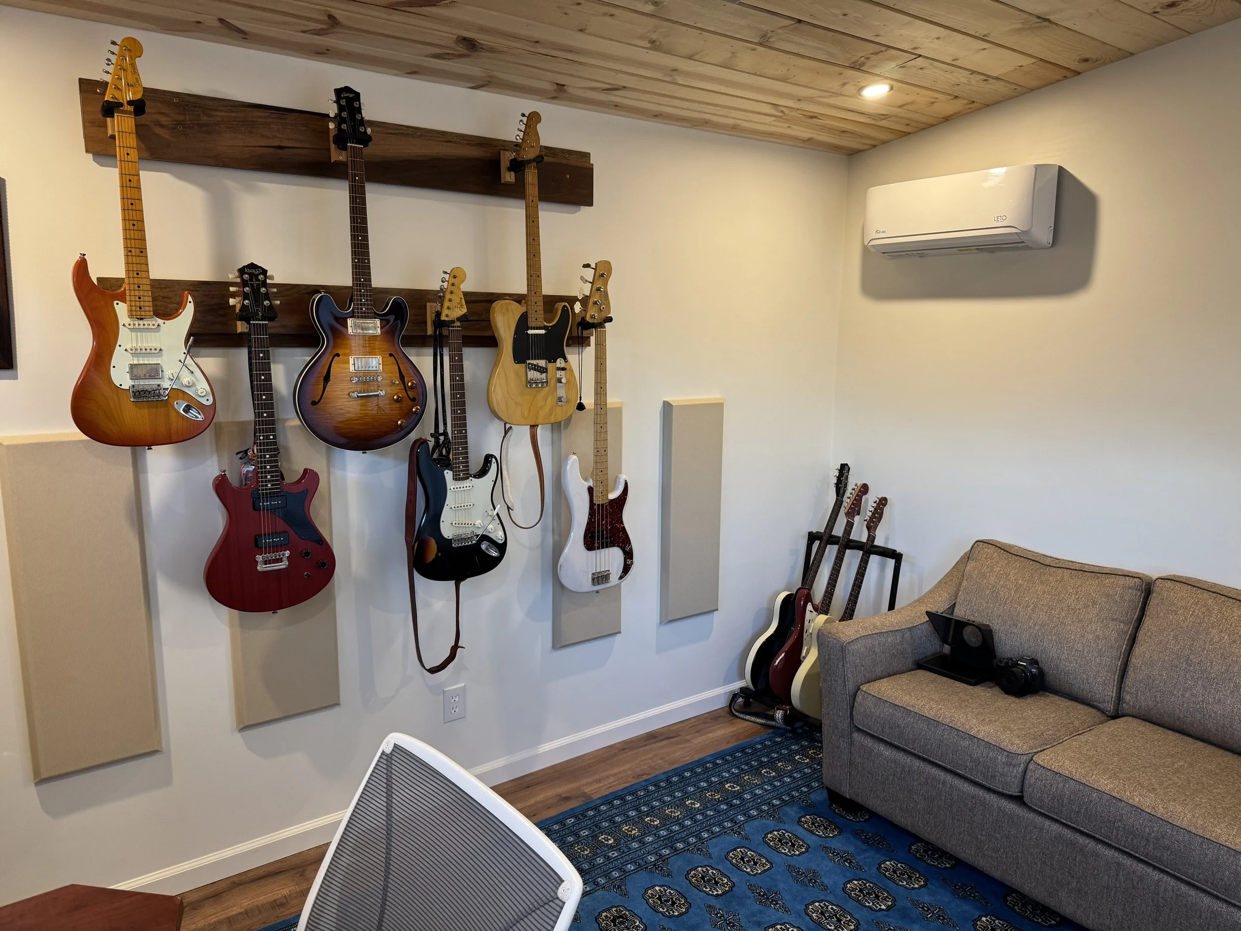 A wall with a wooden ceiling and a white wall with beige acoustic panels, decorated with multiple electric guitars hanging on hooks, a sofa, a blue patterned rug, a camera, and a white chair.