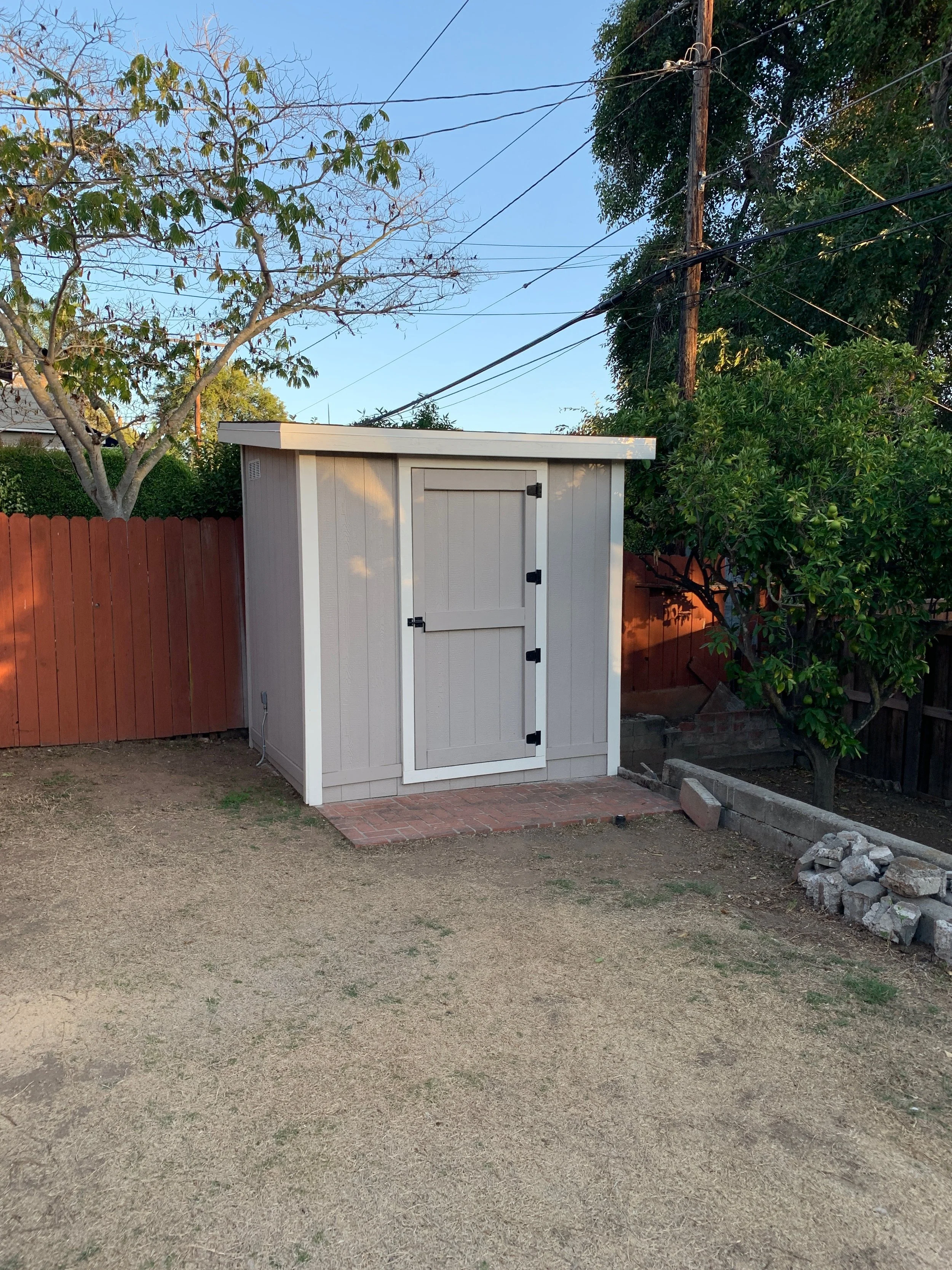 A small white wooden shed with a door, situated on a brick platform in a backyard, with a wooden fence, trees, and utility lines in the background, and a patchy dirt yard in the foreground.