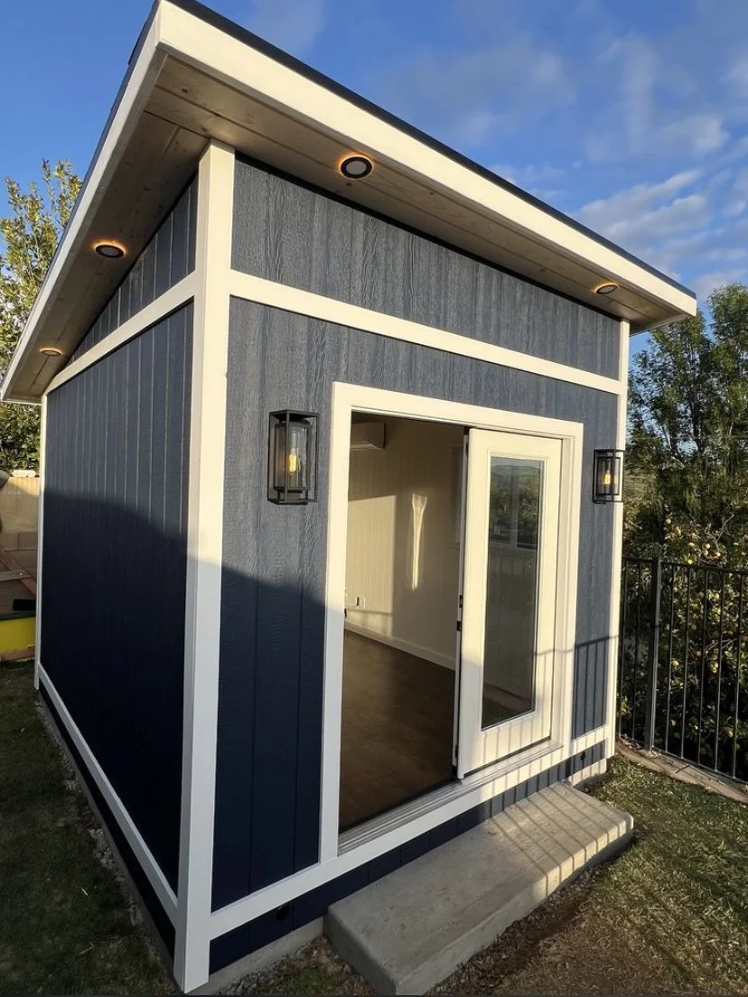 Small modern shed with navy blue siding, white trim, glass door, and black wall-mounted lights, situated outdoors on a concrete step with a backyard fence and trees in the background.