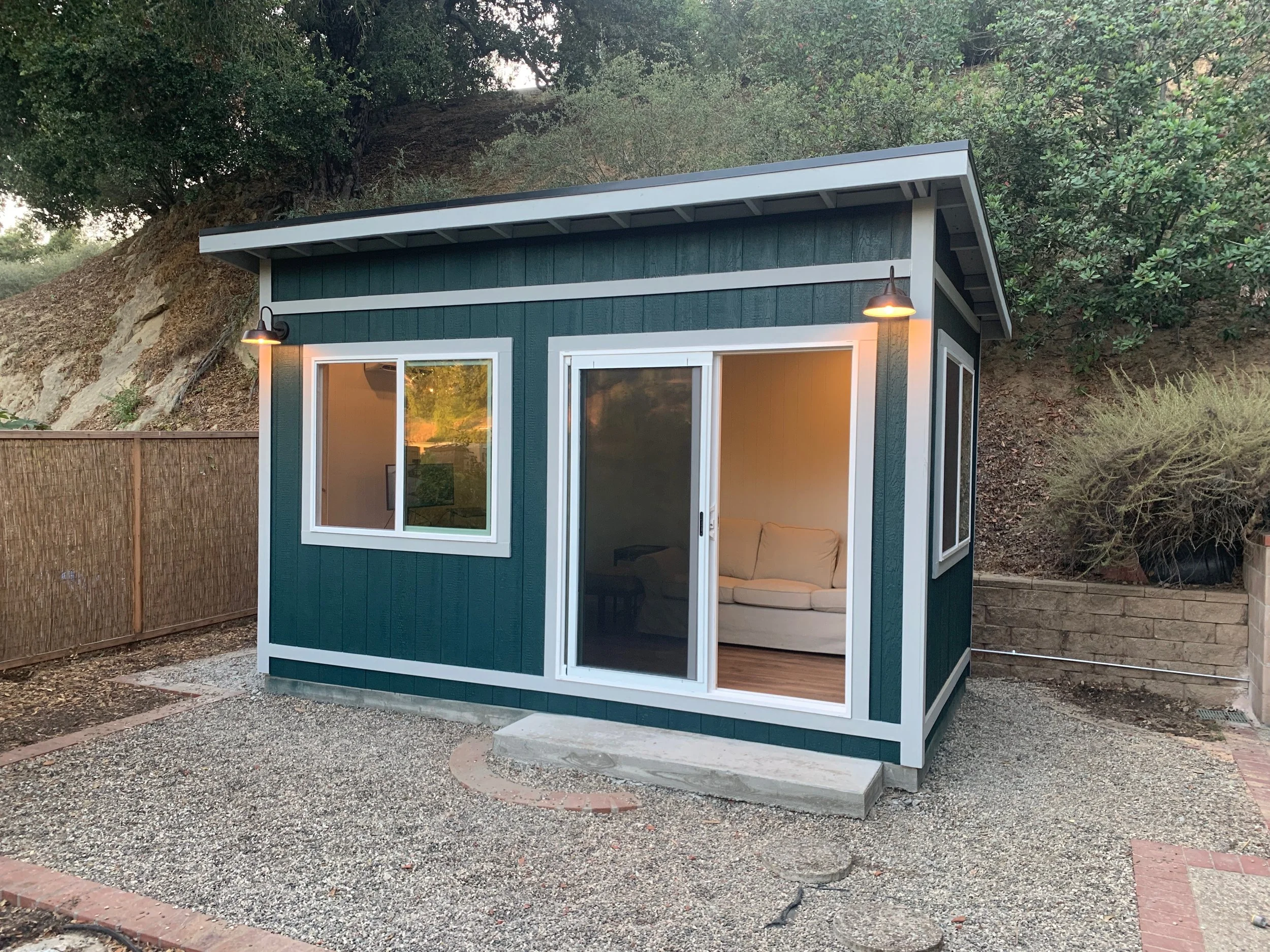 A small, modern backyard shed painted in dark teal with white trim, featuring sliding glass doors and a window, with two external light fixtures above the doors, situated on a gravel patch surrounded by a curved brick border, against a backdrop of tr