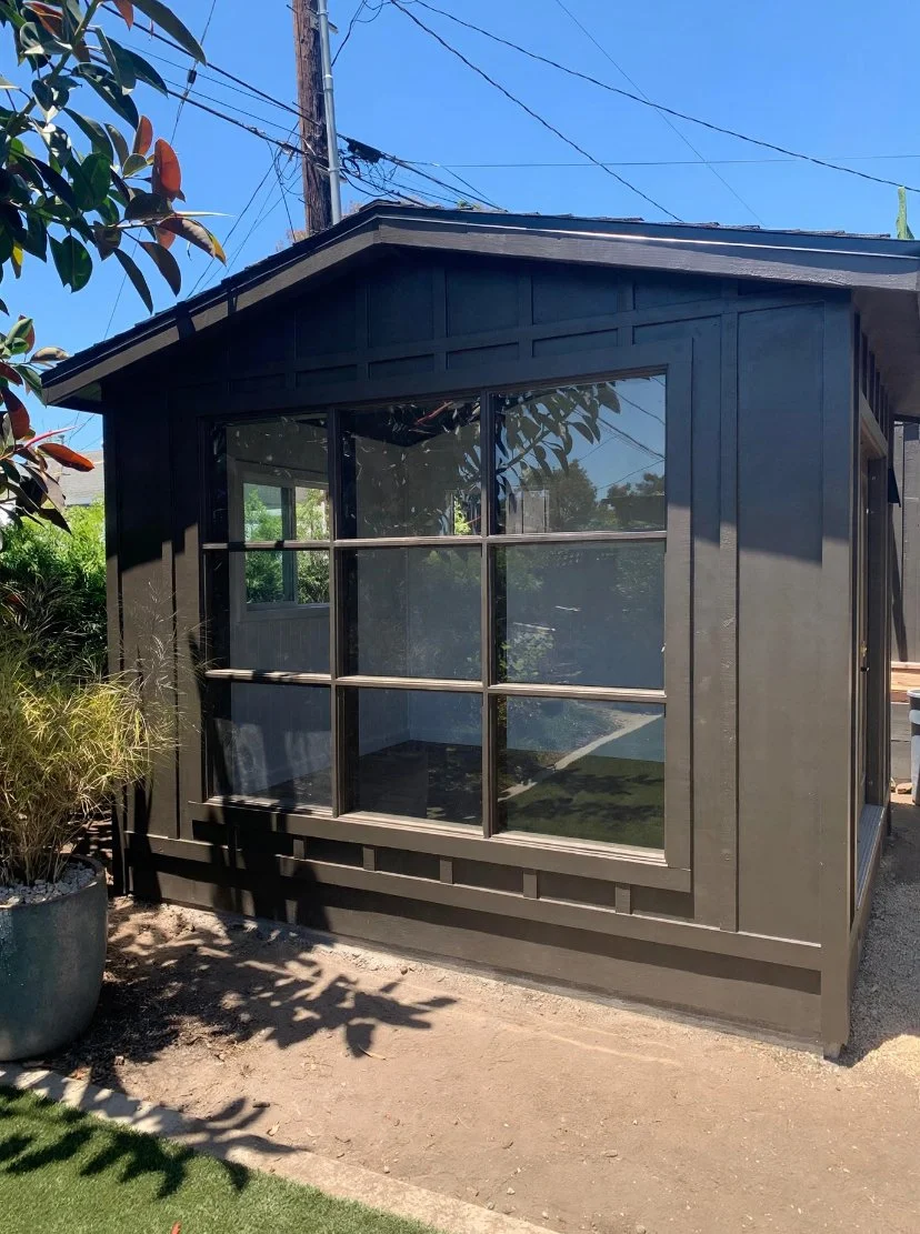 Small black house or shed with large windows, surrounded by plants and trees, under a clear blue sky.