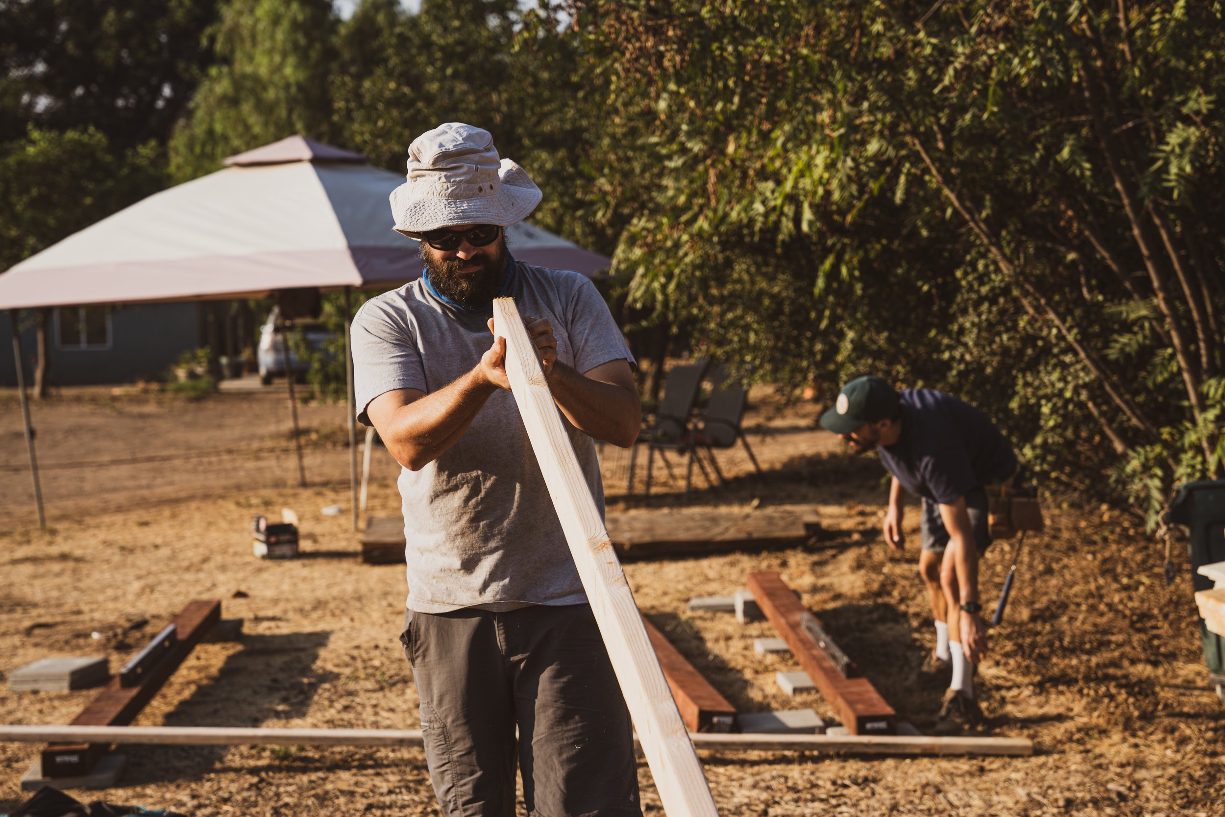 Two men working outdoors on a construction project, with one man holding a wooden plank and the other using a tool on the ground. There is a white canopy tent in the background, surrounded by trees.
