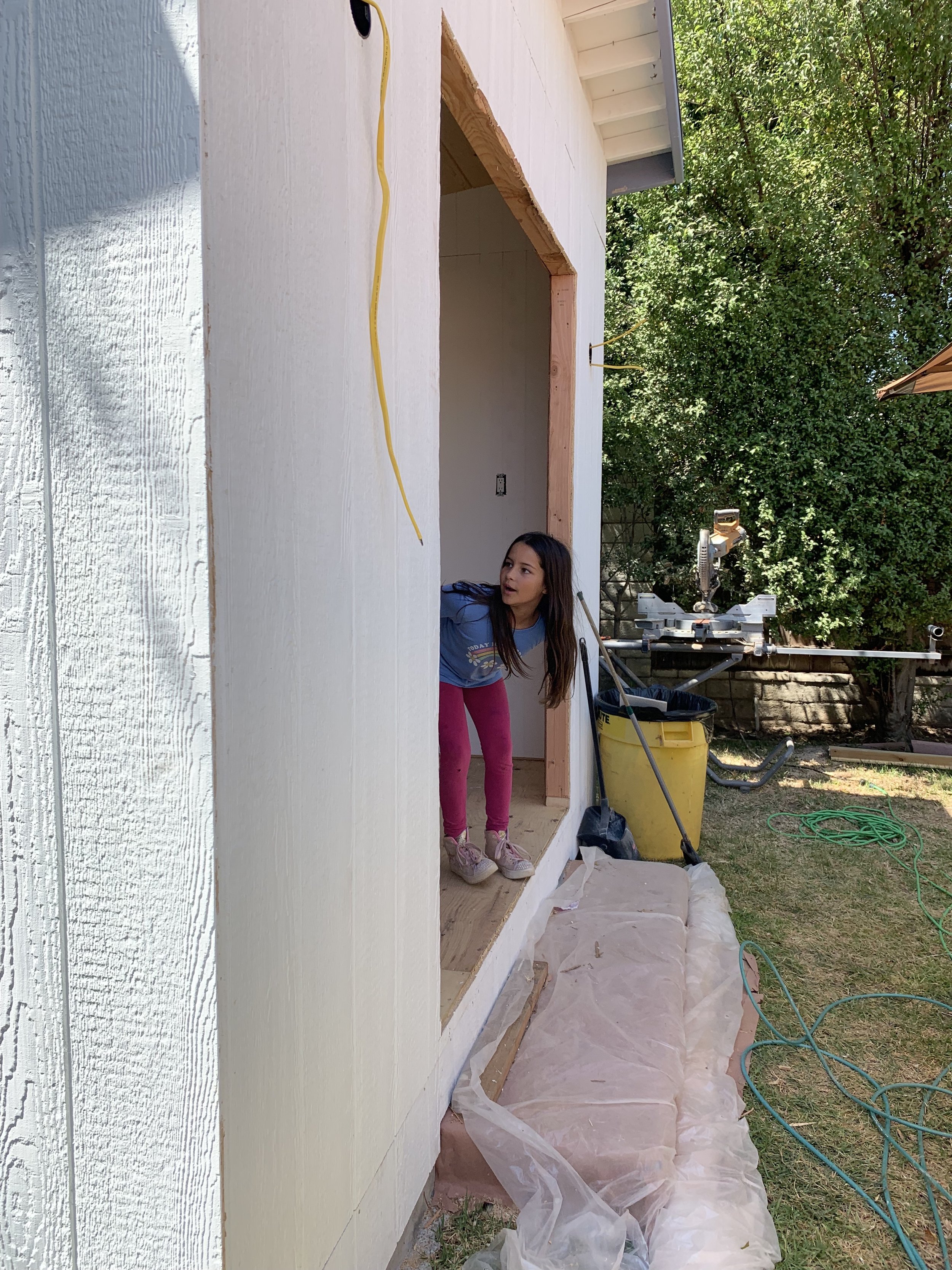 Child peeking out of a doorway of a partially constructed shed with a yellow extension cord hanging. Construction materials and tools visible outside.