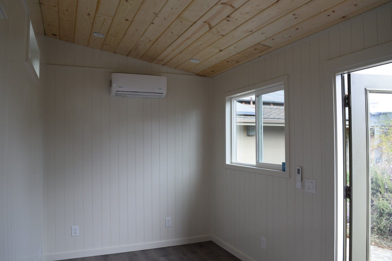 Empty room with white paneled walls, a window, a door, a wall-mounted air conditioner, and wood-paneled ceiling.