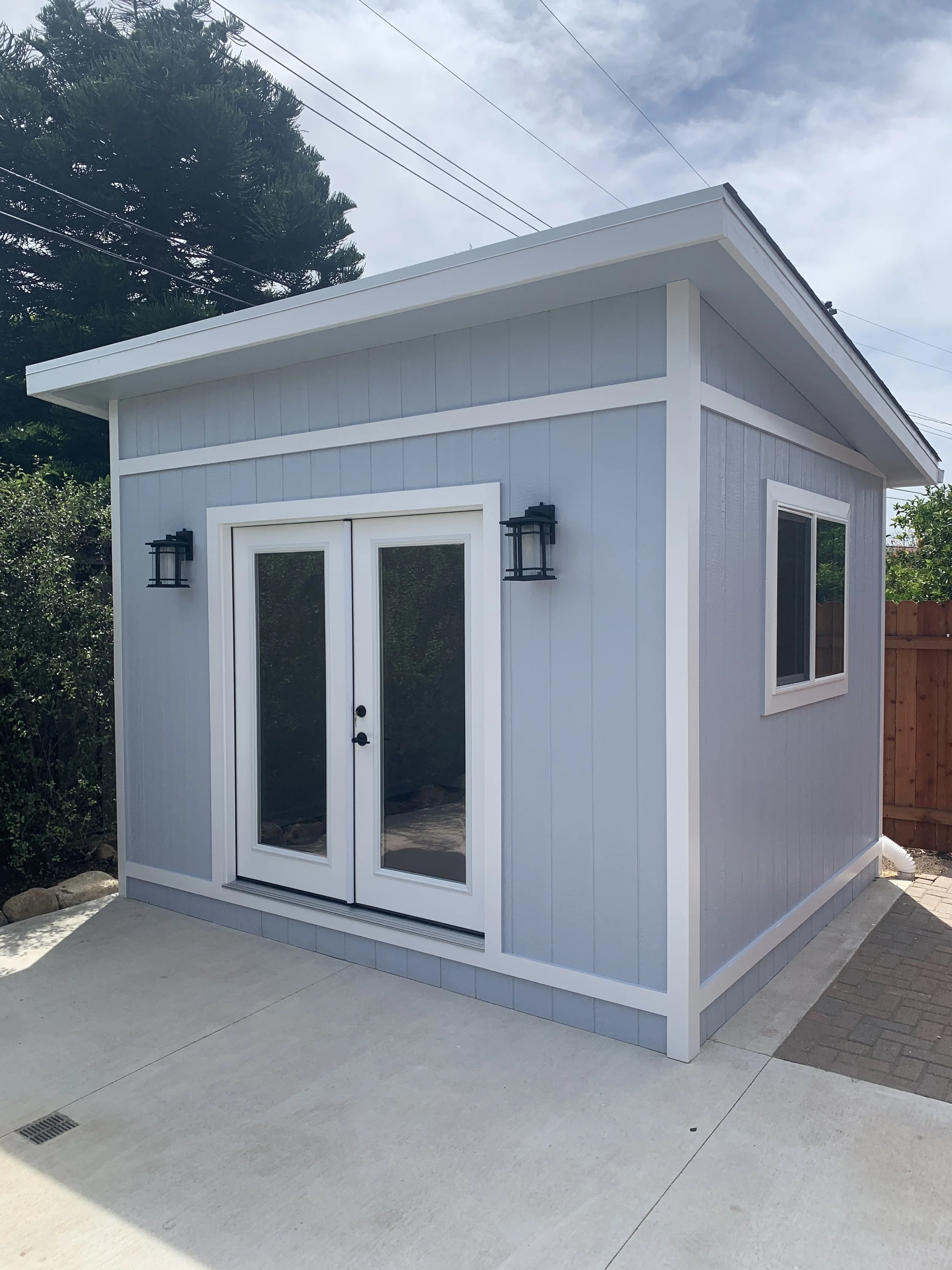 Small modern outdoor shed with white framing, double glass doors, a window, wall-mounted black lanterns, and a concrete patio in front, with trees and utility wires in the background.