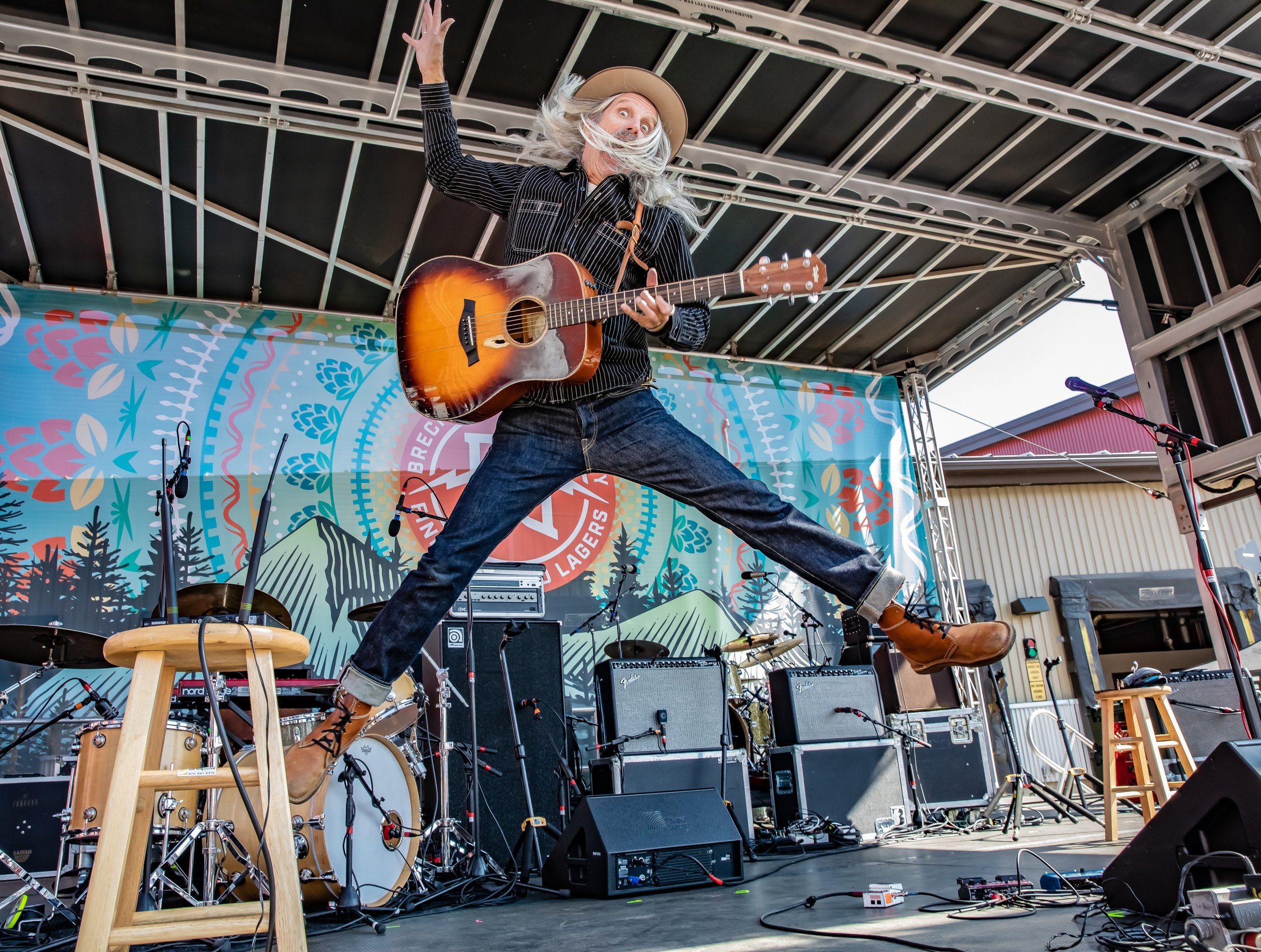 A man with long gray hair, wearing a tan hat, black striped shirt, jeans, and brown boots, jumps in the air while playing an acoustic guitar on a stage at an outdoor event.