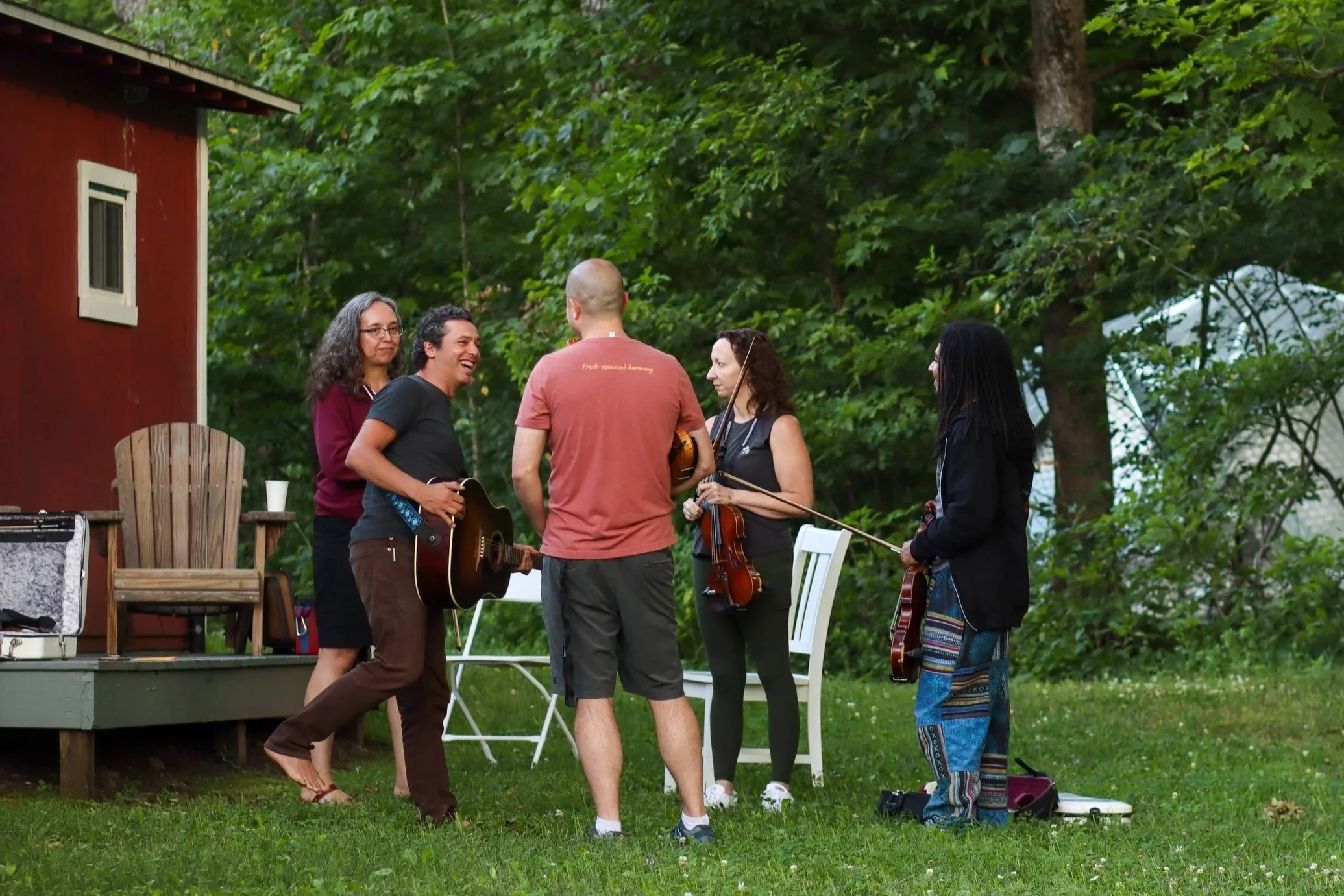 Group of five people standing outdoors on a grassy area with trees in the background, holding musical instruments and talking, near a small wooden deck with chairs and a red building.