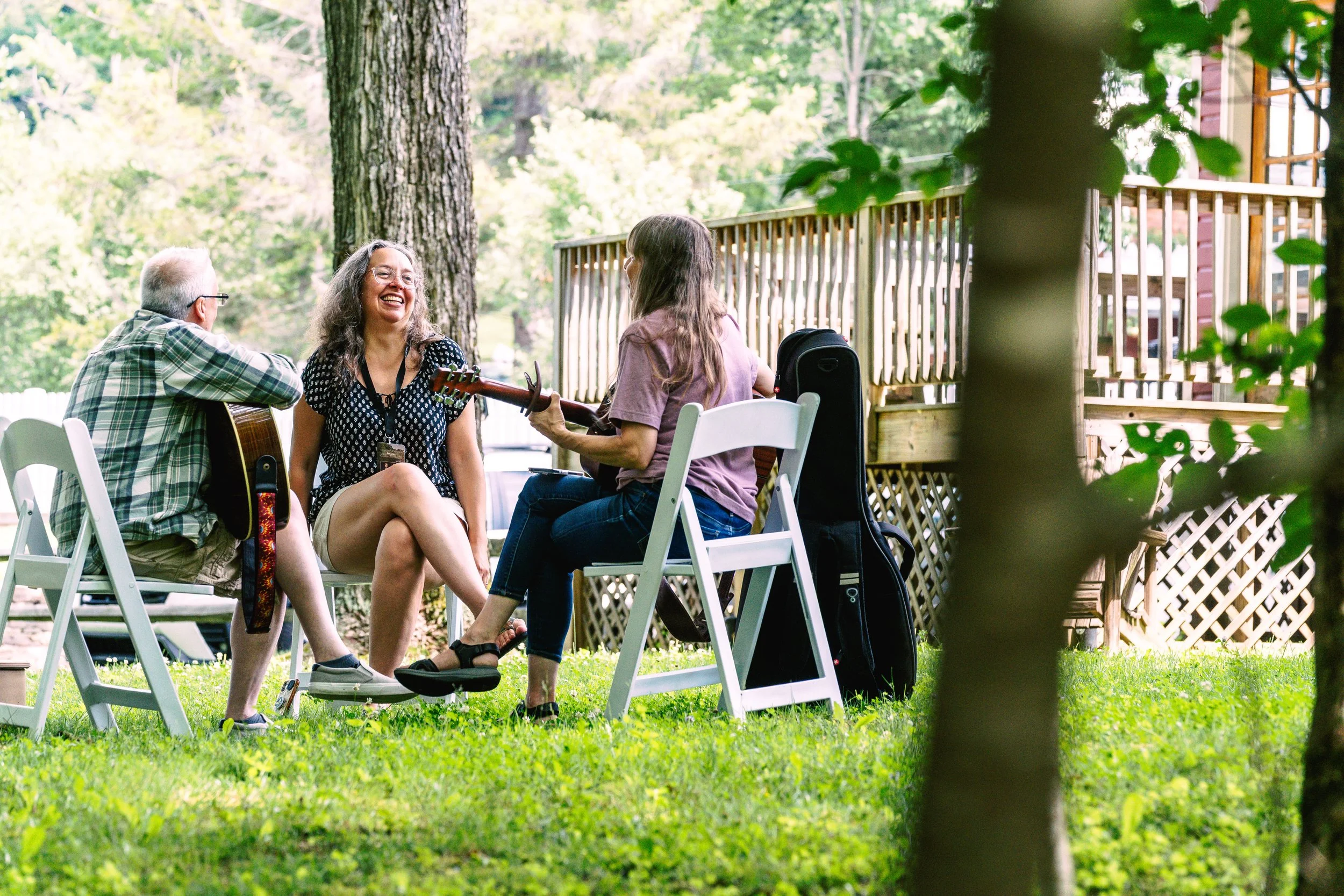 Three people sitting outdoors on chairs, enjoying music and conversation in a backyard with trees and a wooden deck in the background.