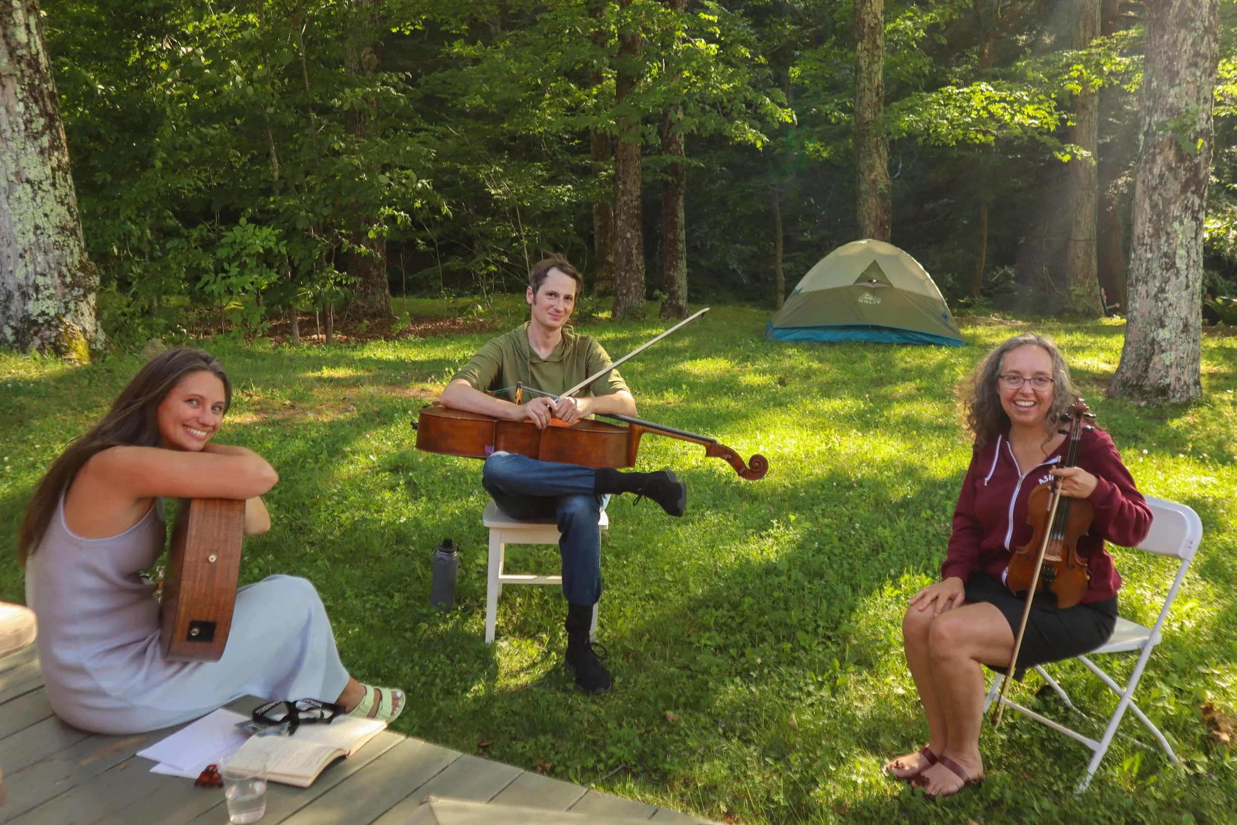 Three friends playing music outdoors in a forest clearing with a tent in the background. One woman is sitting on a wooden deck with a notebook and camera, smiling. The man in the middle is sitting on a white chair, playing a cello, and the woman on t