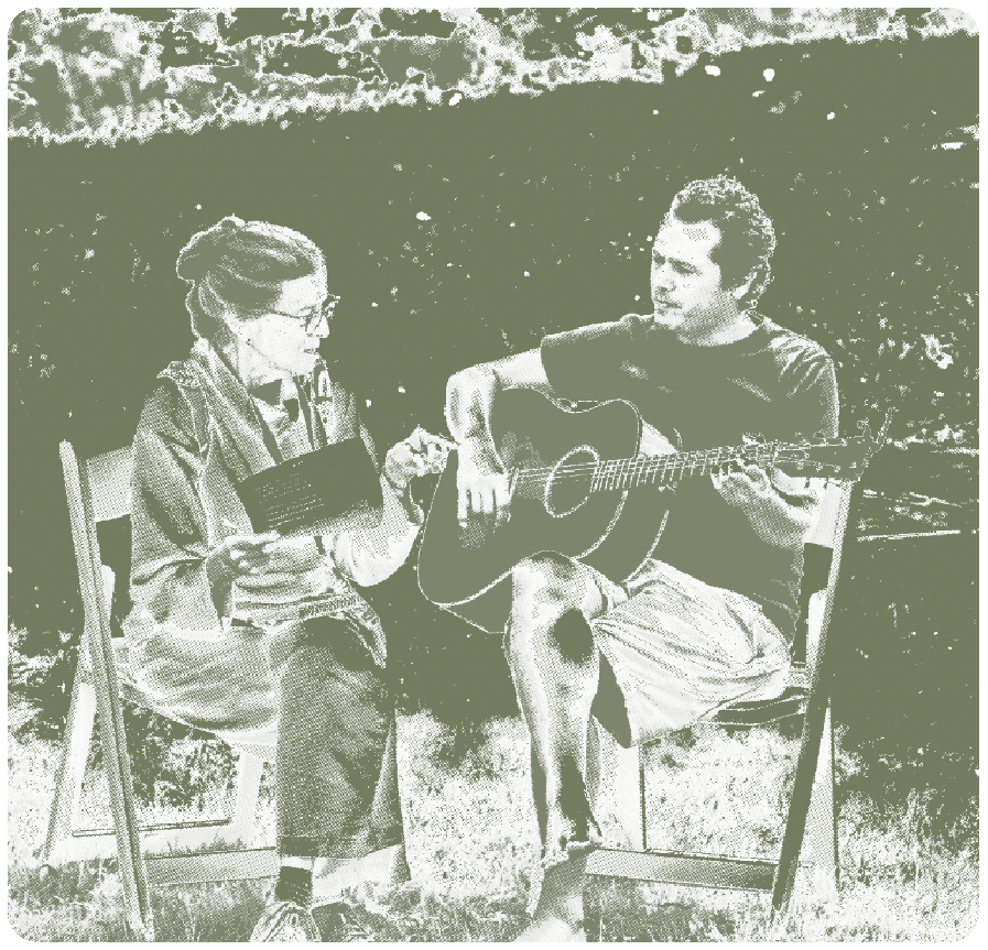 A woman and a man sitting outdoors near the water, engaged in playing guitar and reading.