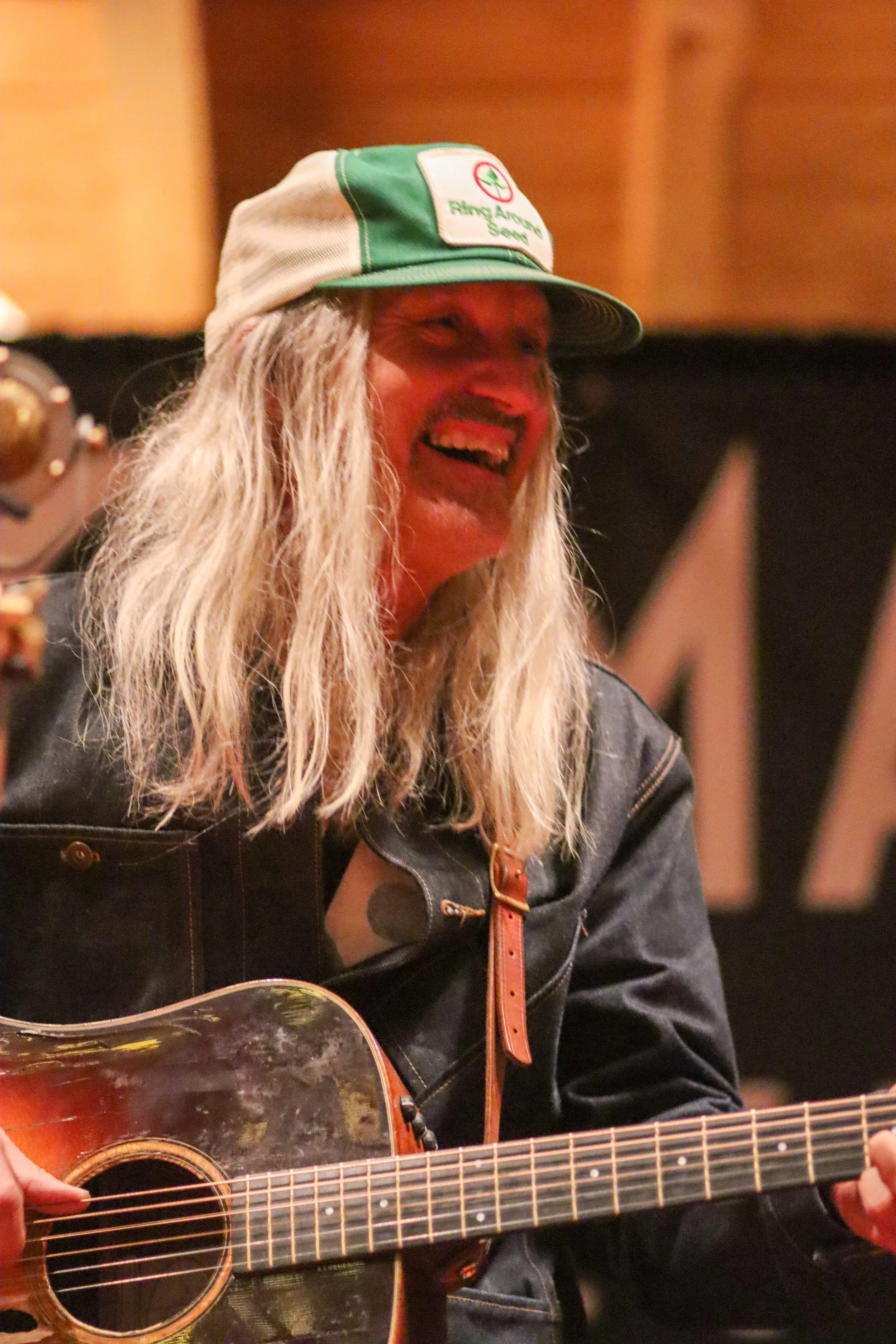 A man with long blond hair wearing a green and white cap and a black denim jacket, playing an acoustic guitar and smiling.