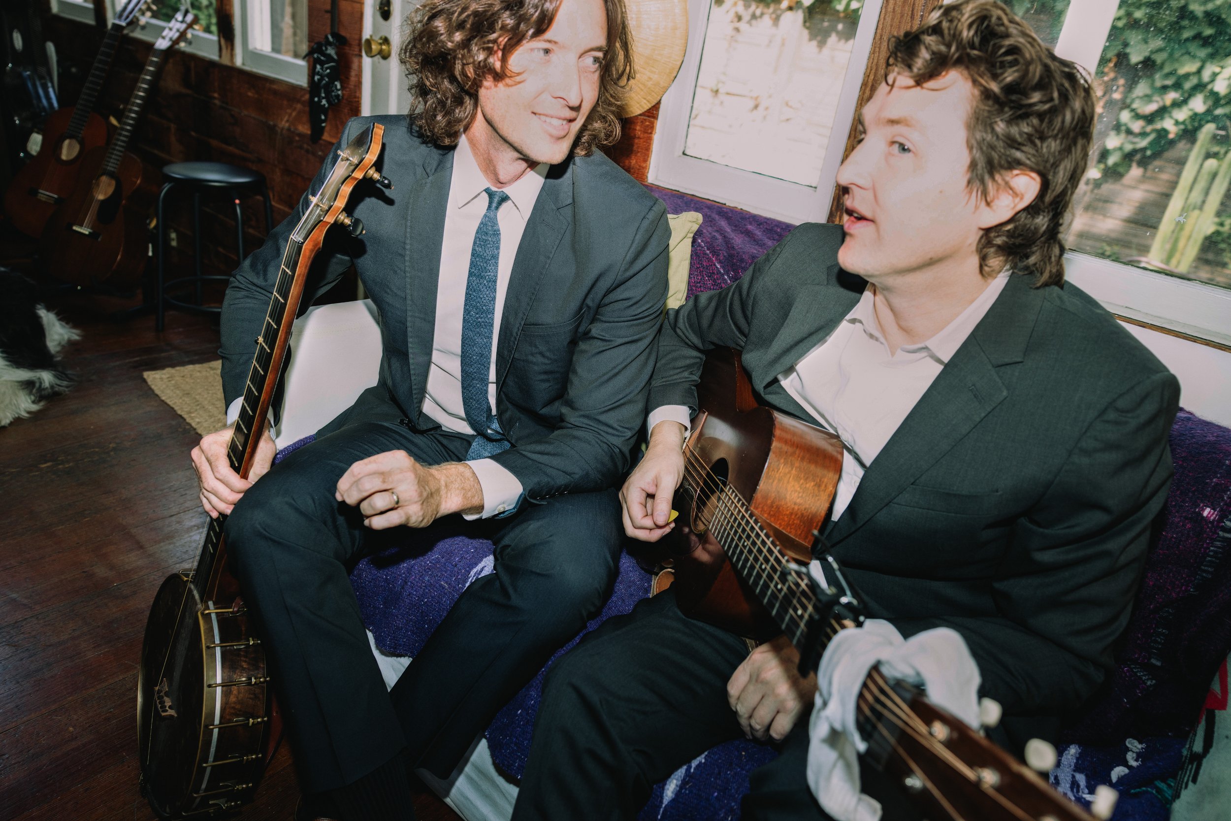 Two men in suits sitting on a couch inside a cozy room, playing guitars and sharing a moment of music and conversation.