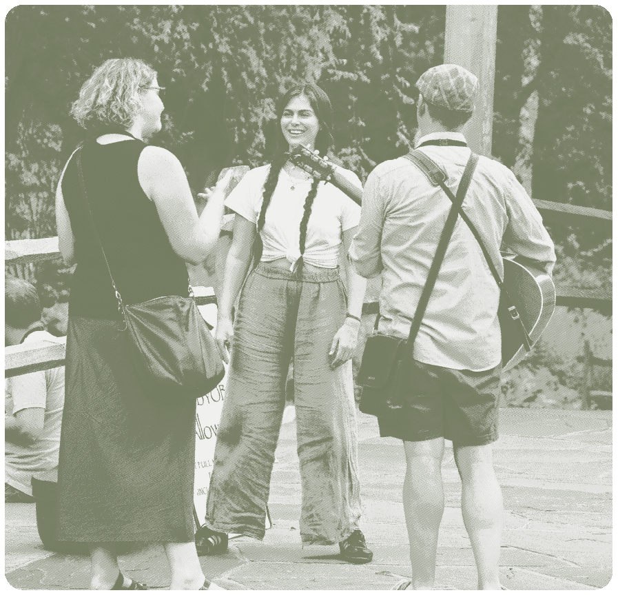 A young woman with long dark hair holding a microphone and smiling, standing between two people, a woman and a man, outdoors with trees in the background. The woman is dressed casually, while the woman and man are dressed casually, with the man holding a guitar.