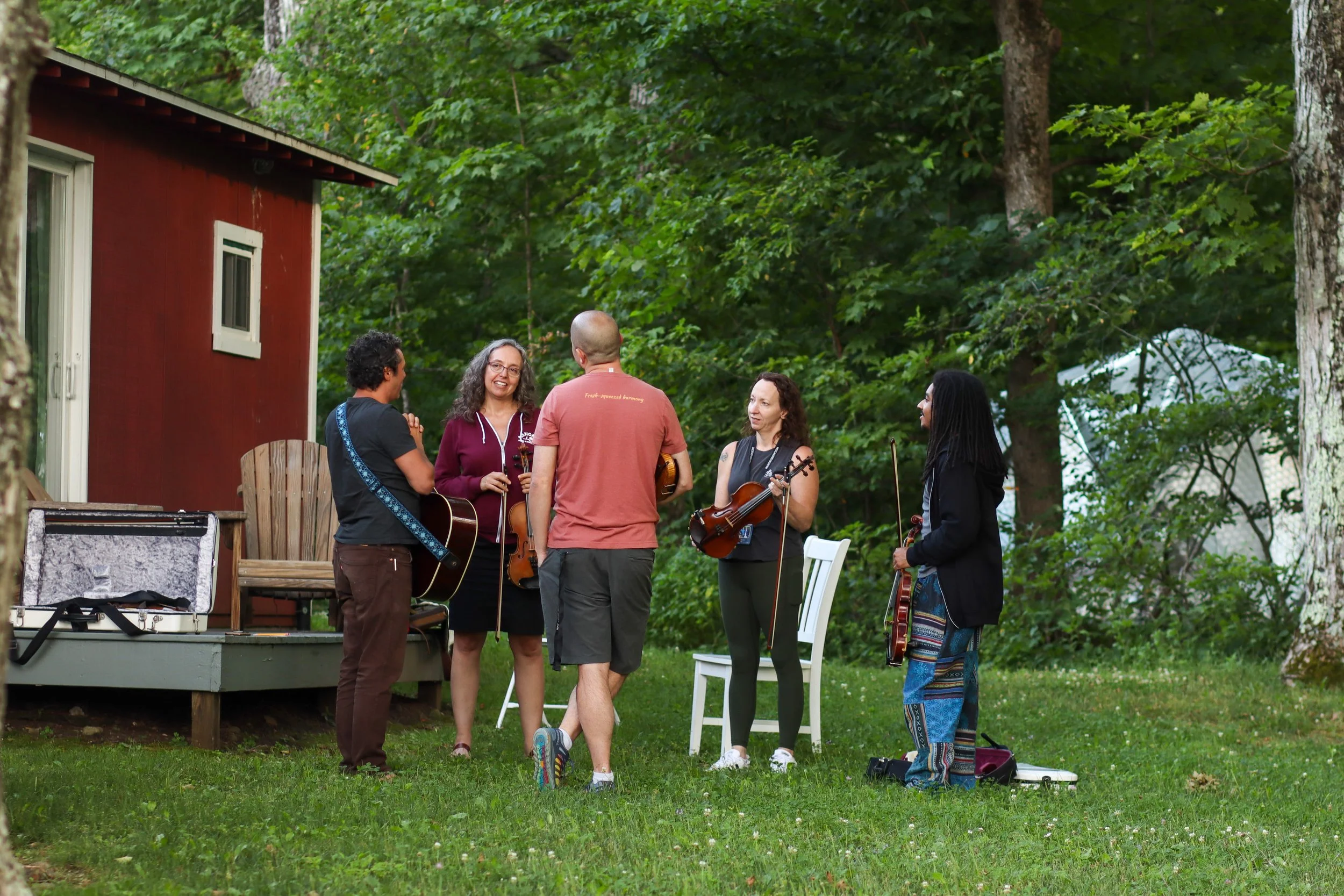 A group of five people gathered outdoors on green grass playing musical instruments and chatting near a red house with a porch and white-framed windows, surrounded by trees.