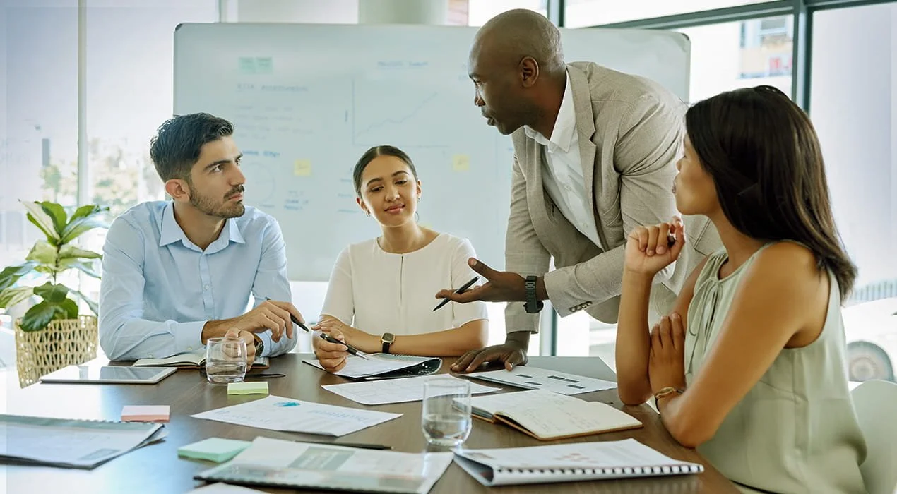 Business meeting in a modern conference room with four diverse individuals, one man speaking and three women listening, surrounded by documents, notebooks, and glasses of water.