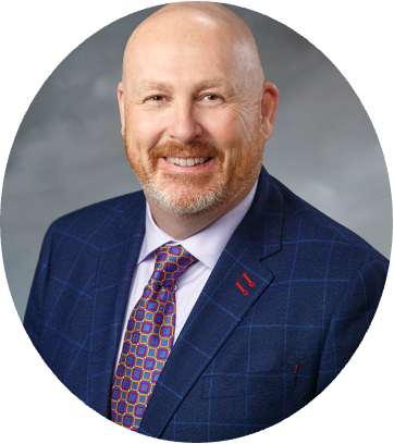 Professional headshot of a bald man with a beard, wearing a blue checked suit, a white shirt, and a colorful patterned tie, smiling against a gray background.