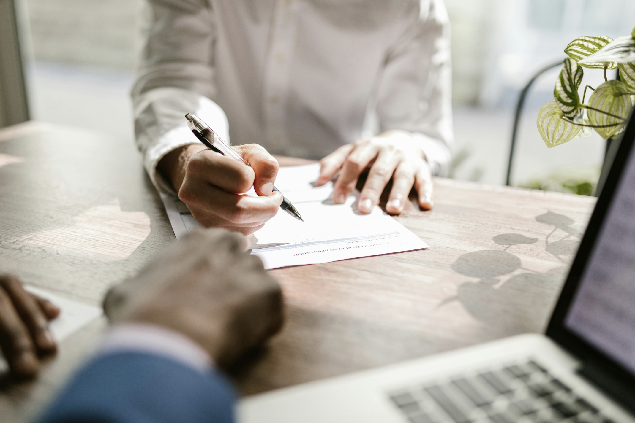 Person signing a document with a pen while seated at a wooden table near a window, with a laptop partially visible in the foreground and a potted plant on the side.