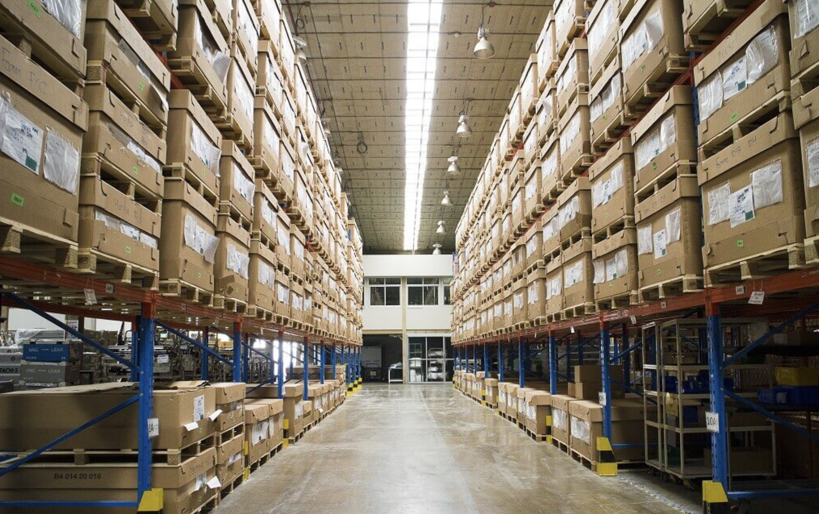 Warehouse aisle with tall shelves filled with cardboard boxes, some labeled, and a smooth concrete floor leading to a white door at the far end.