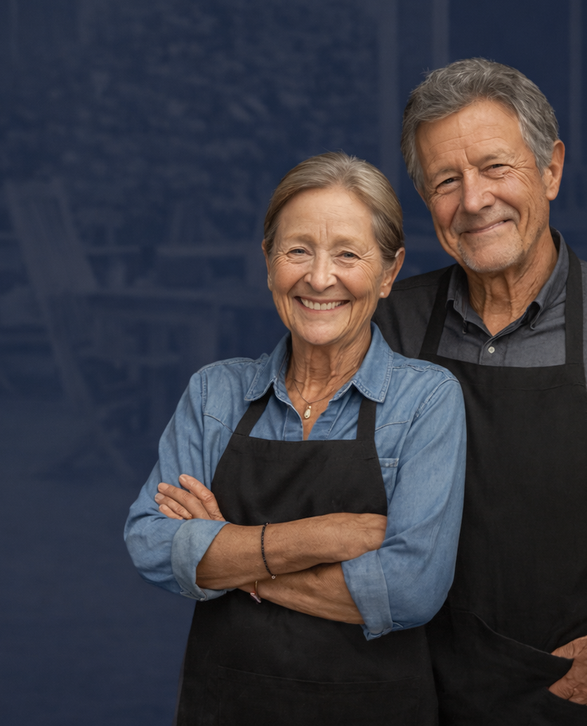An elderly couple smiling while wearing black aprons, standing close together in a friendly pose.