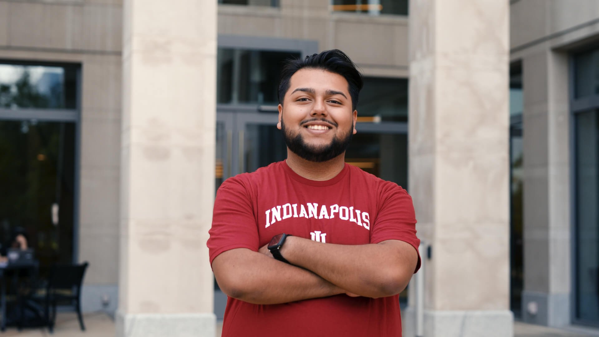 Smiling person in a red "Indianapolis IU" shirt, standing with arms crossed in front of a modern building.