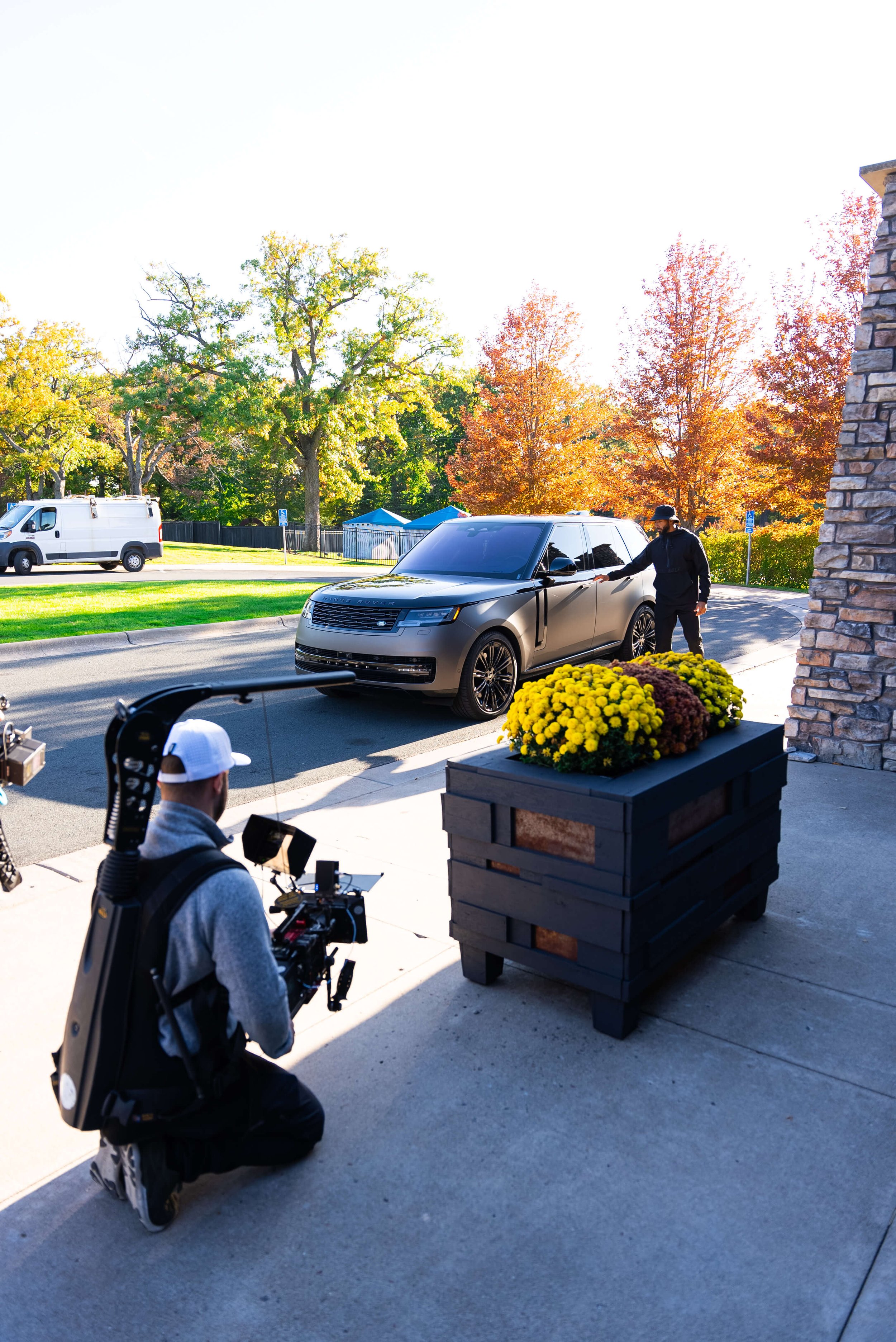 Filmmaker with camera and harness filming matte black SUV near flowers and autumn trees.