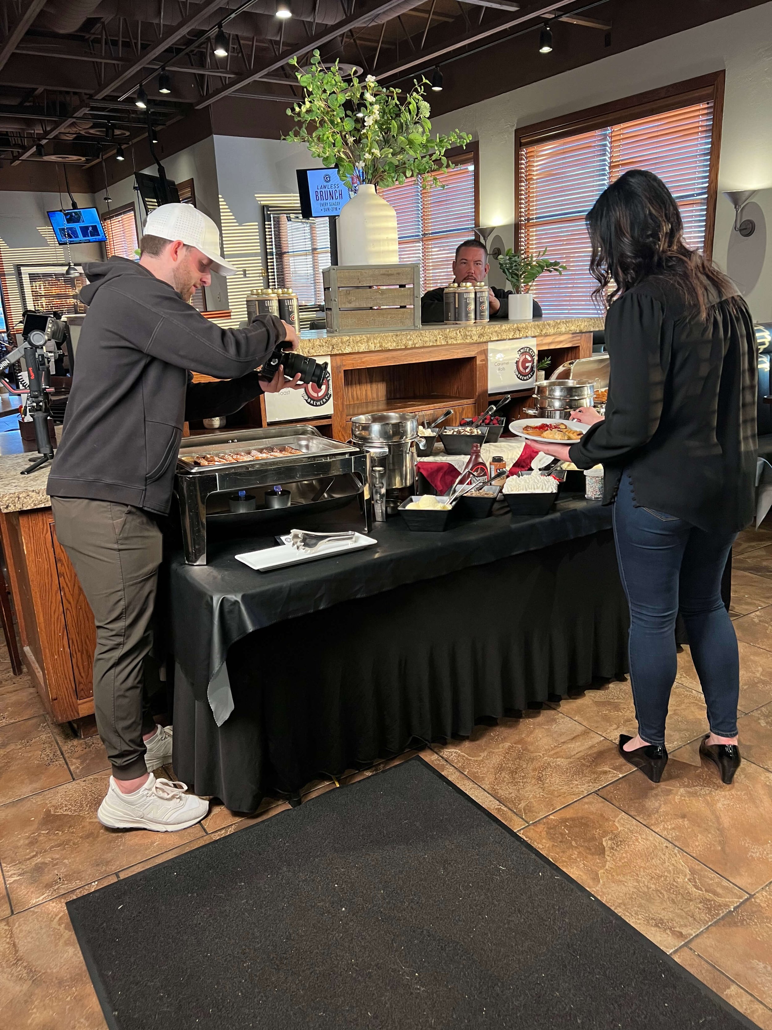 Two people serving themselves food from a buffet table at a restaurant, with a chef in the background and a plant decoration above.