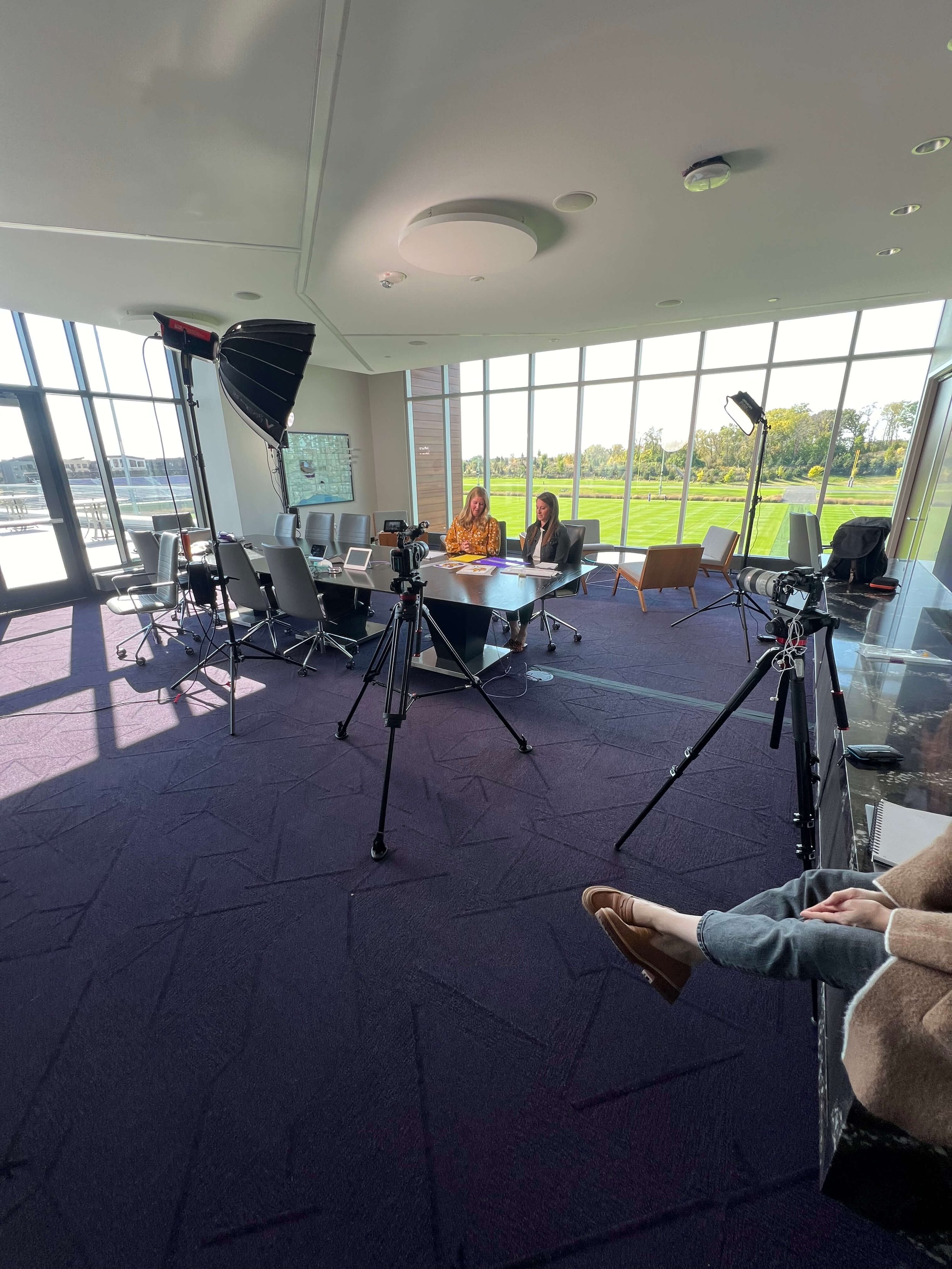Office setup with video recording equipment, two people at a table, large windows, and lighting equipment.