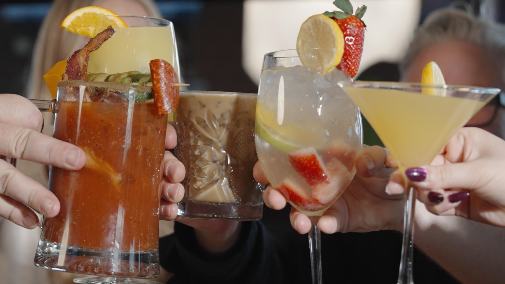 People holding various cocktails, including a Bloody Mary, iced coffee, sangria, and a martini, adorned with fruit garnishes.