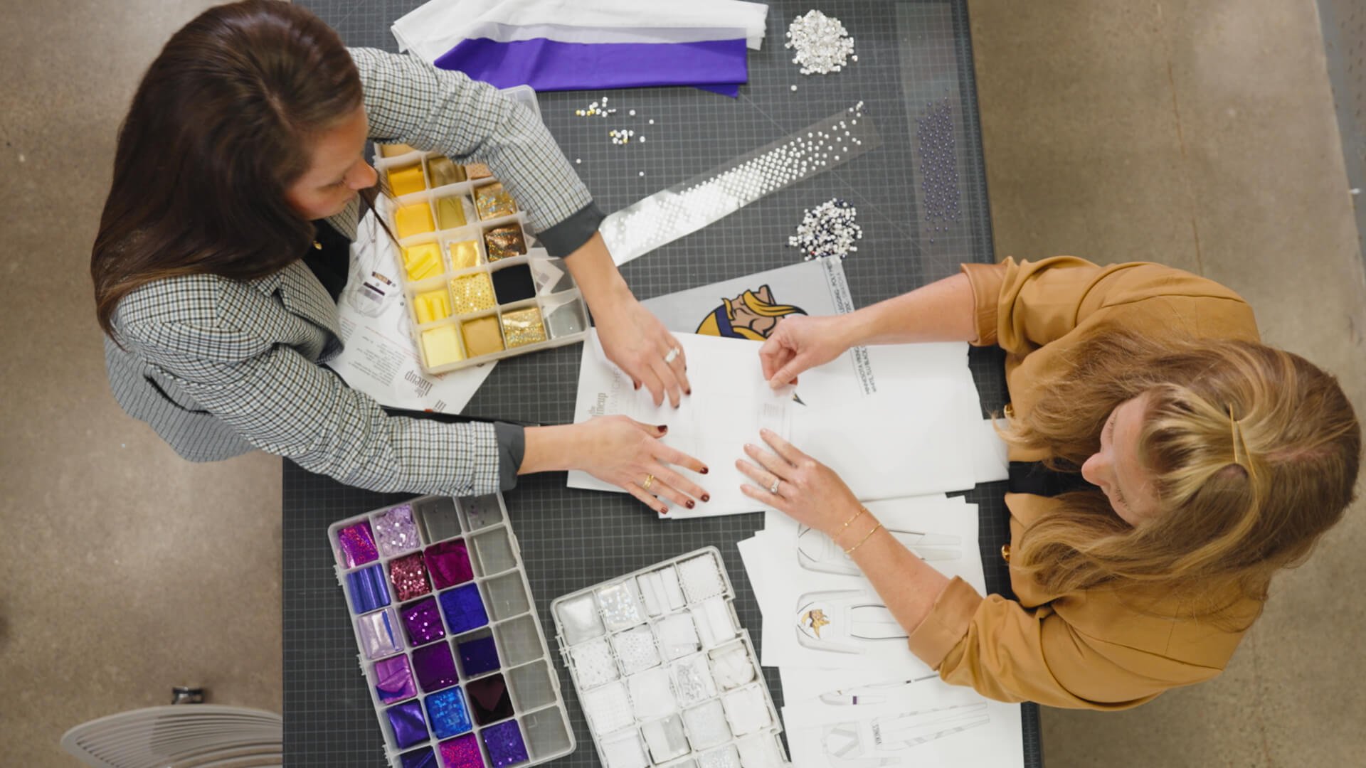 Two women working on design projects at a table, with fabric samples and sketches. Various colored and textured materials are organized in trays.