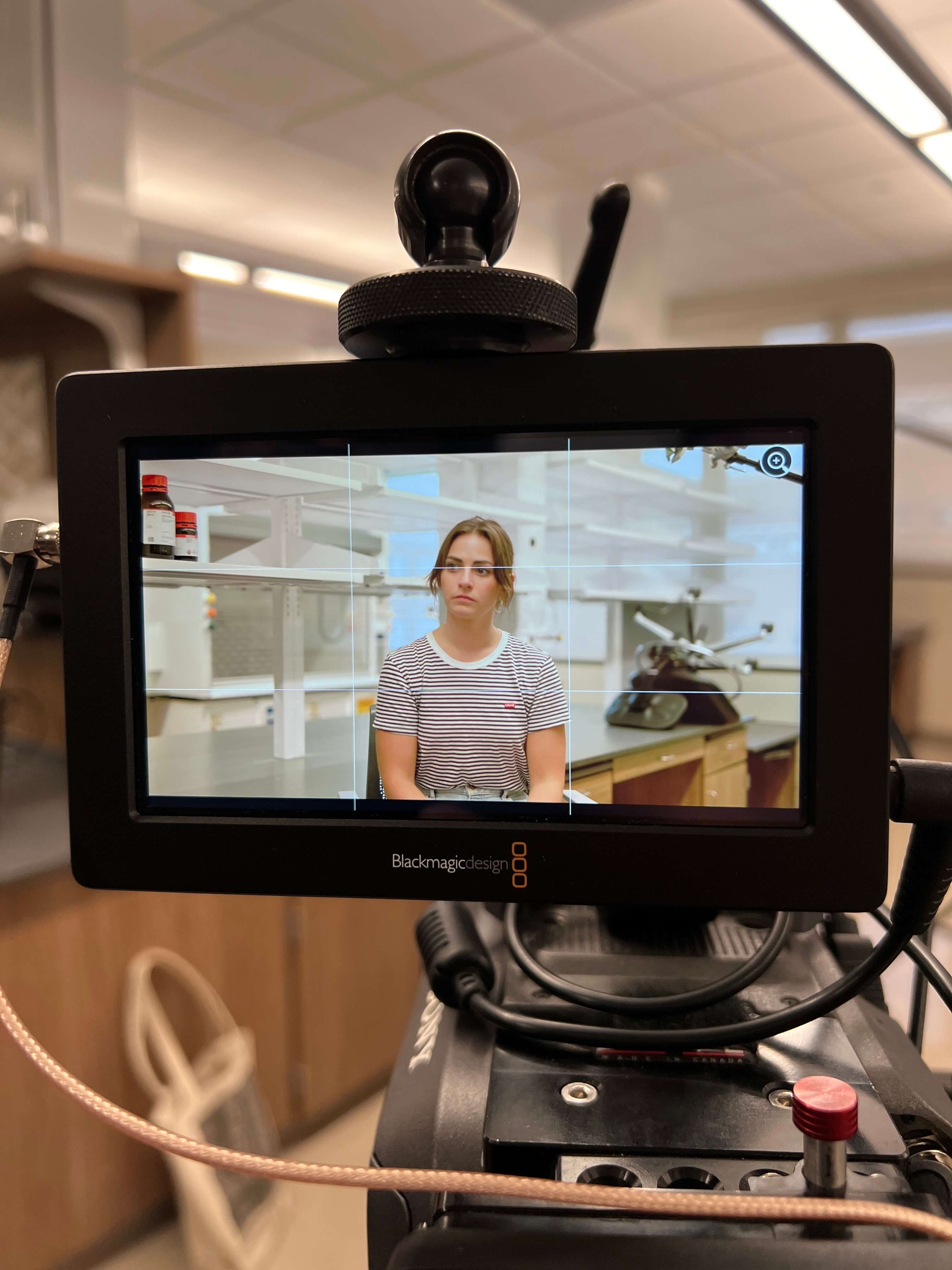 A video camera screen displaying a person with brown hair wearing a striped shirt, sitting in a laboratory environment. Equipment and shelves with bottles are visible in the background.