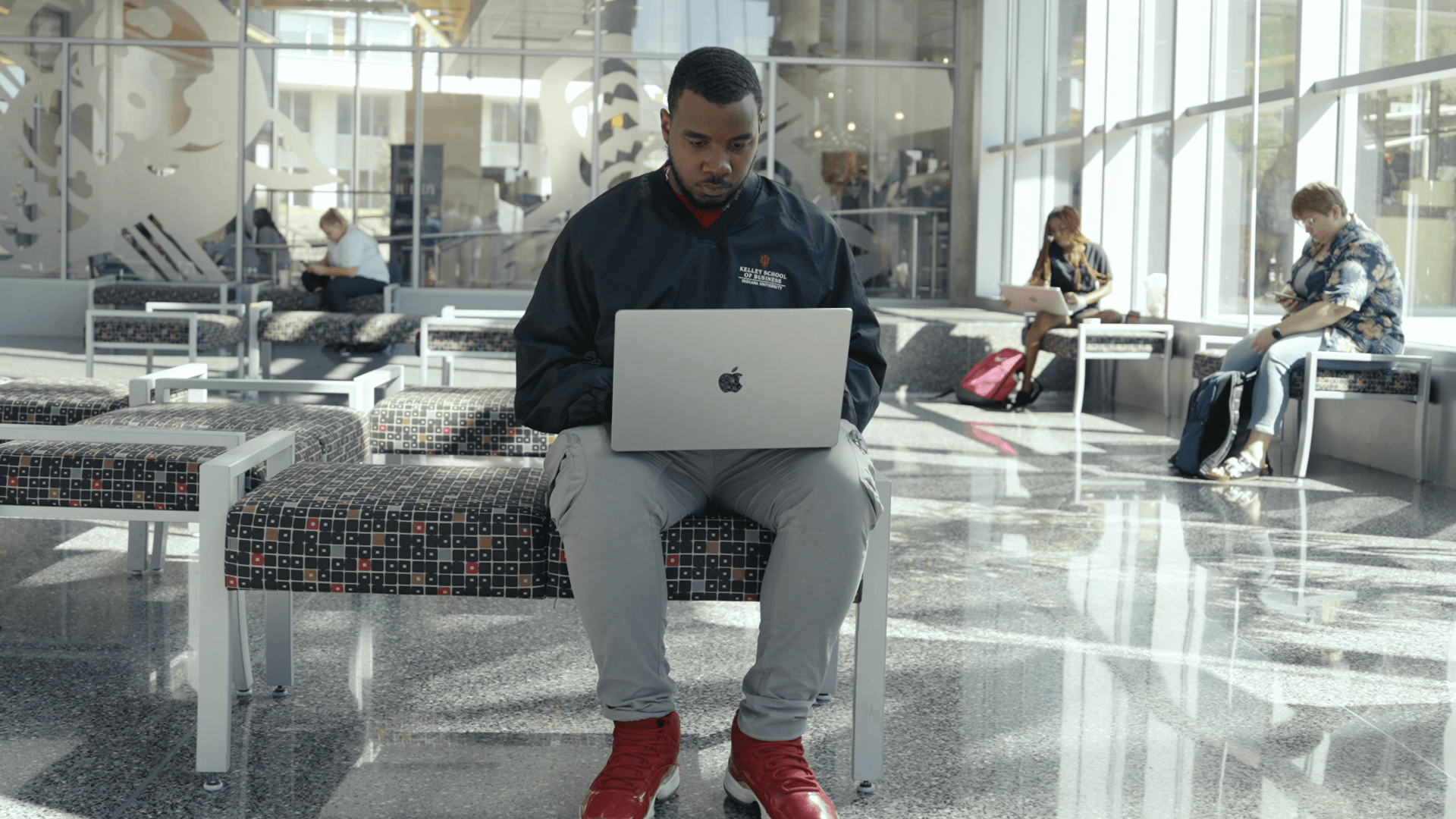 Man sitting on a bench using a laptop, with other people in the background, inside a modern building with large windows.